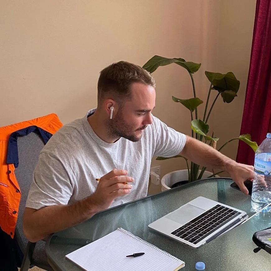 A man seated at a glass table working on a laptop, with a notepad, water bottle, and wireless earbuds, indoors with a potted plant and red curtain in the background.