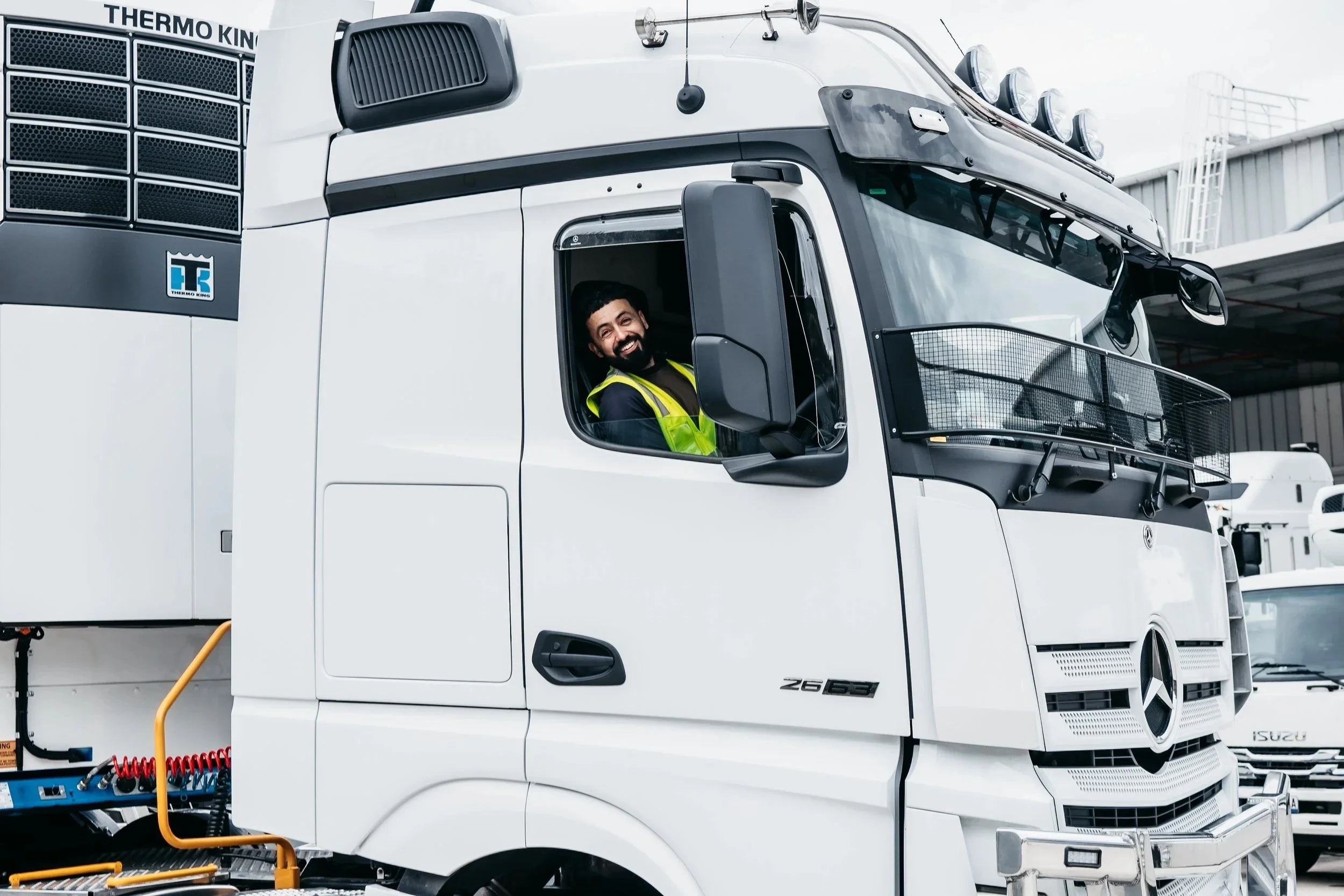 A smiling man in a yellow safety vest sitting in the driver’s seat of a large white semi-truck, parked in an industrial area.