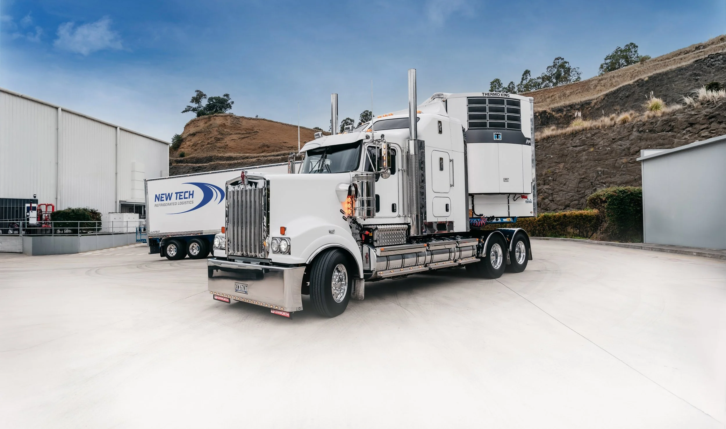 A large white semi-truck with a sleeper cab and refrigerator logistics branding on the side, parked on a concrete lot with industrial buildings and hills in the background.