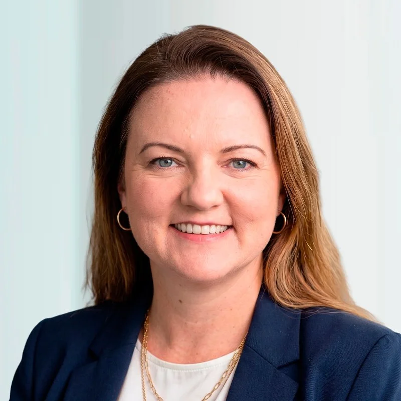 A professional woman with shoulder-length brown hair, wearing a navy blazer, white top, gold jewelry, and hoop earrings, smiling at the camera.