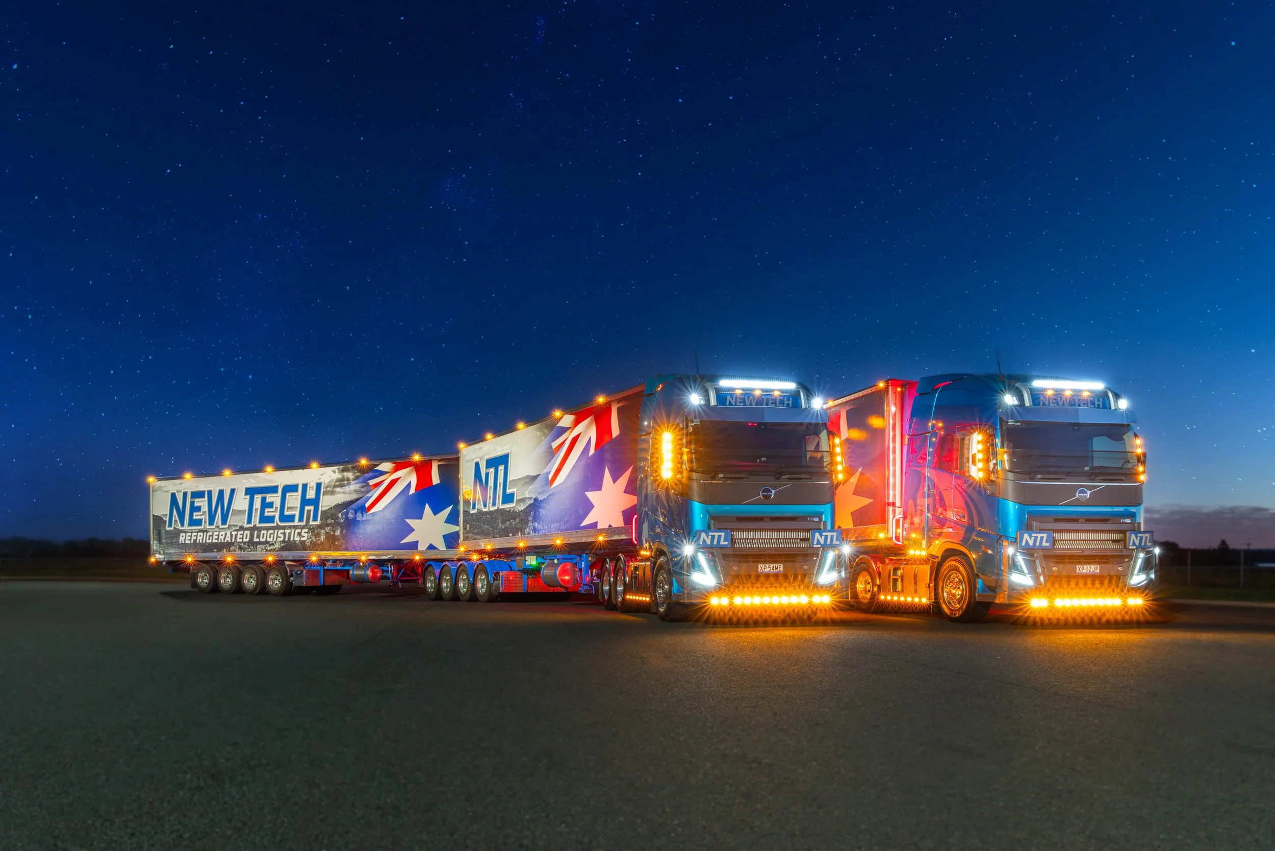 Two large semi-trucks with colorful, illuminated designs and company branding parked side by side on an open road at night, under a starry sky.