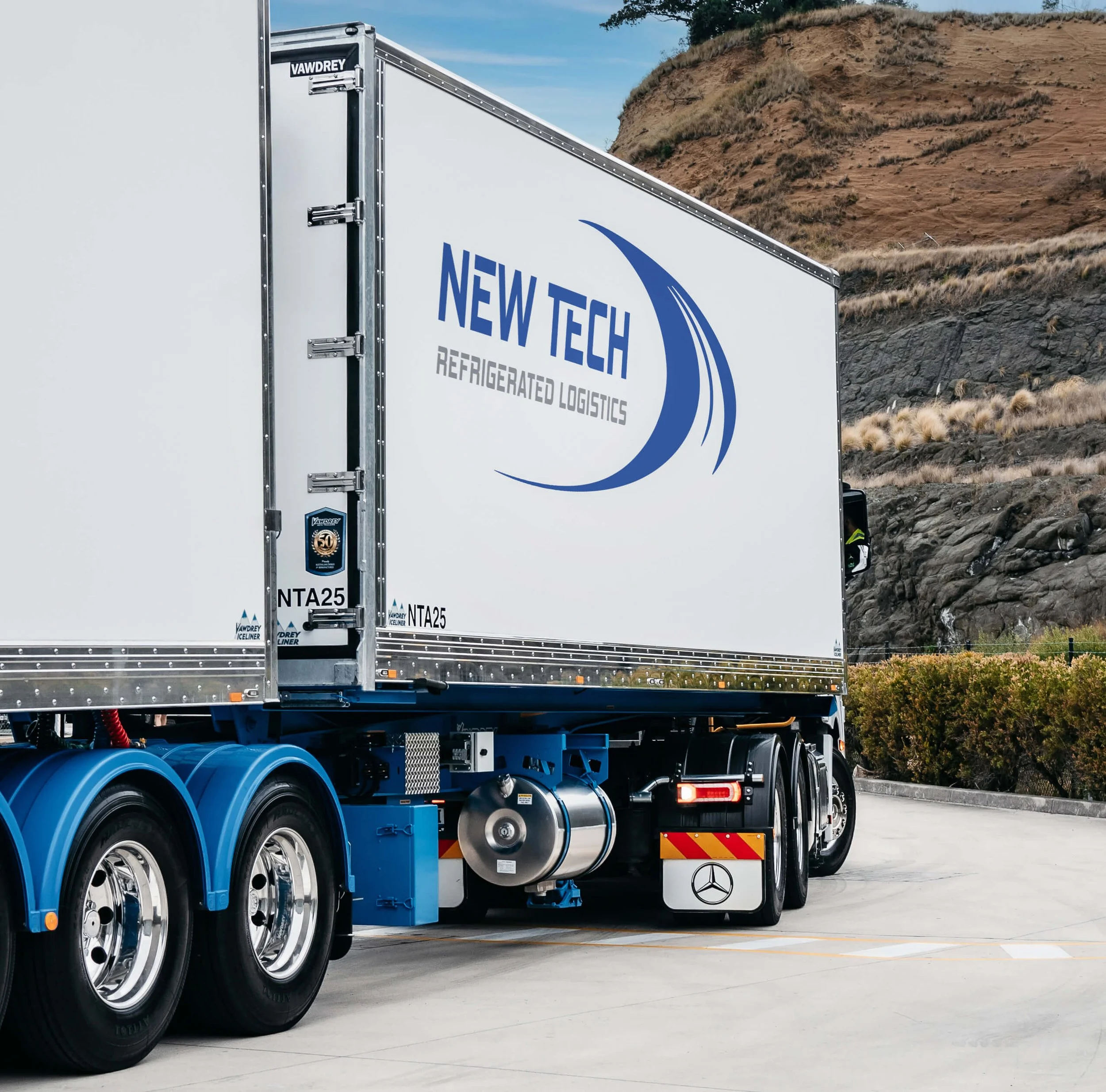 A refrigerated logistics truck parked outside with mountainous terrain in the background. The truck has the words 'NEW TECH REFRIGERATED LOGISTICS' on its side and appears to be part of a fleet, with blue wheels and a Mercedes-Benz logo visible.