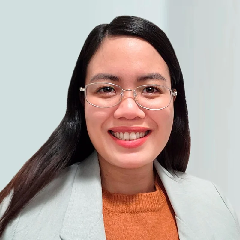 A woman with long dark hair, glasses, and a bright smile, wearing a light gray blazer over an orange sweater.