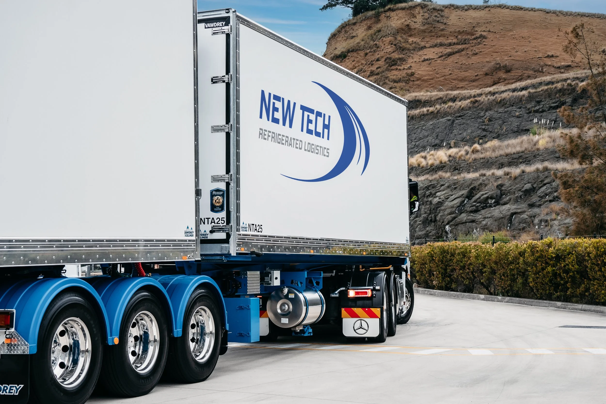 A large white freight truck with blue wheels and branding that reads "New Tech Refrigerated Logistics," parked on a paved surface near a landscaped area with brown shrubbery and a hill in the background.