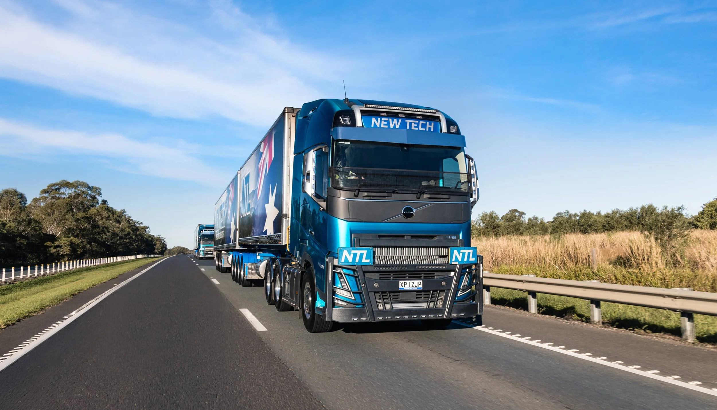 Blue semi-truck with branding, traveling on a highway through a rural area with grass and trees, under a partly cloudy sky.