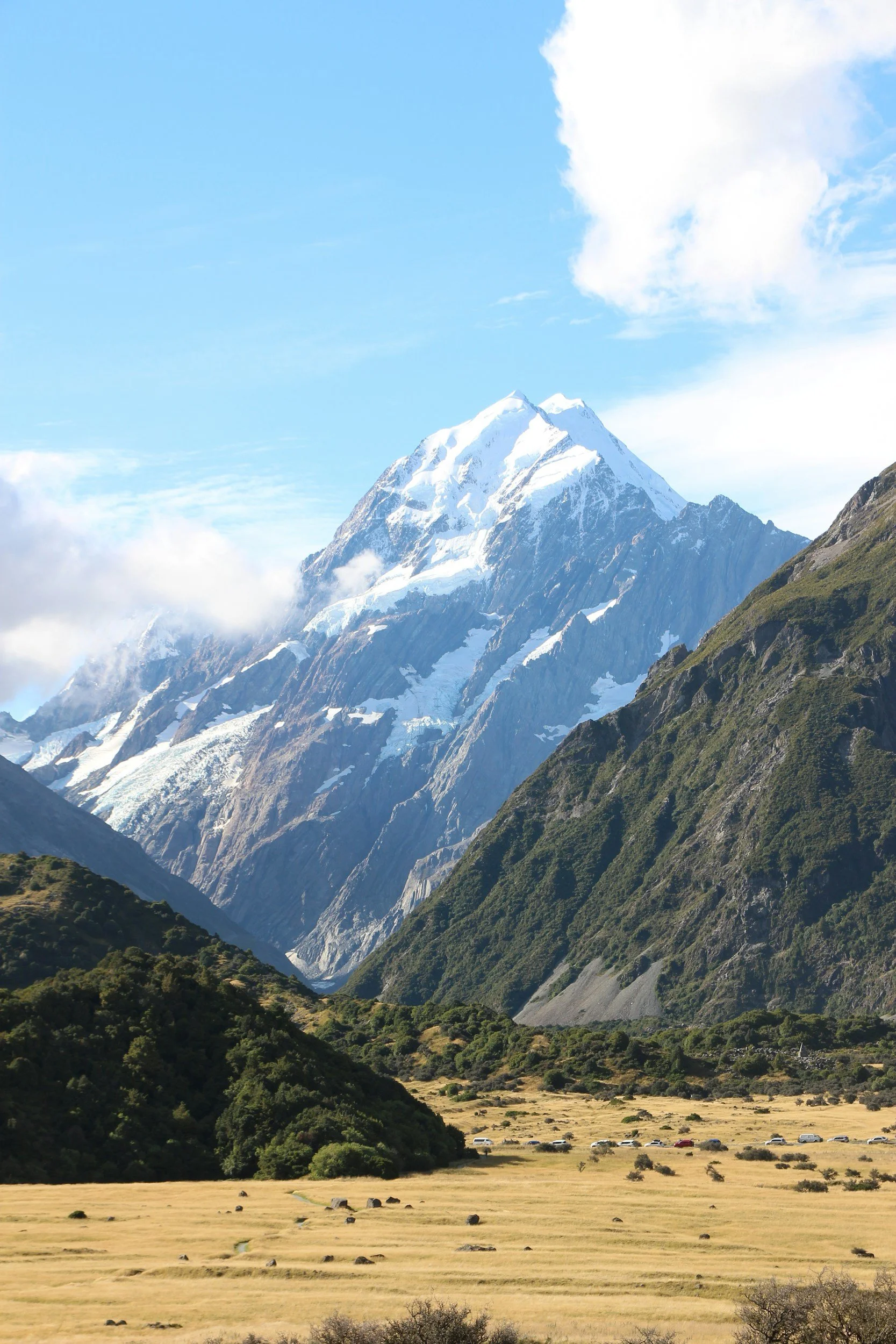 A scenic view of a snow-capped mountain range with green hills in the foreground under a partly cloudy sky.