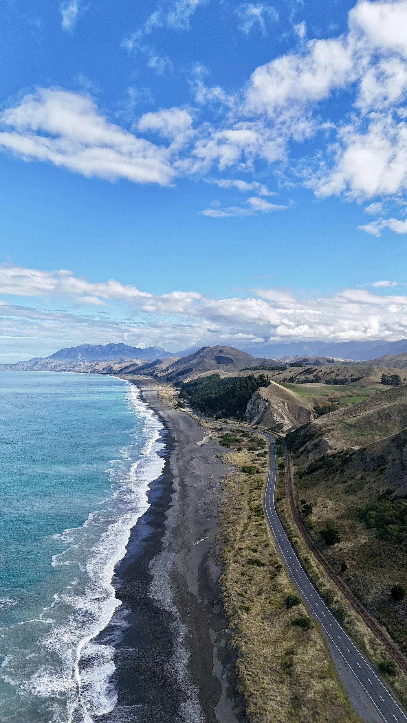 Aerial view of rugged coastline with mountains in the background, ocean on the left, and a winding road parallel to the shore.
