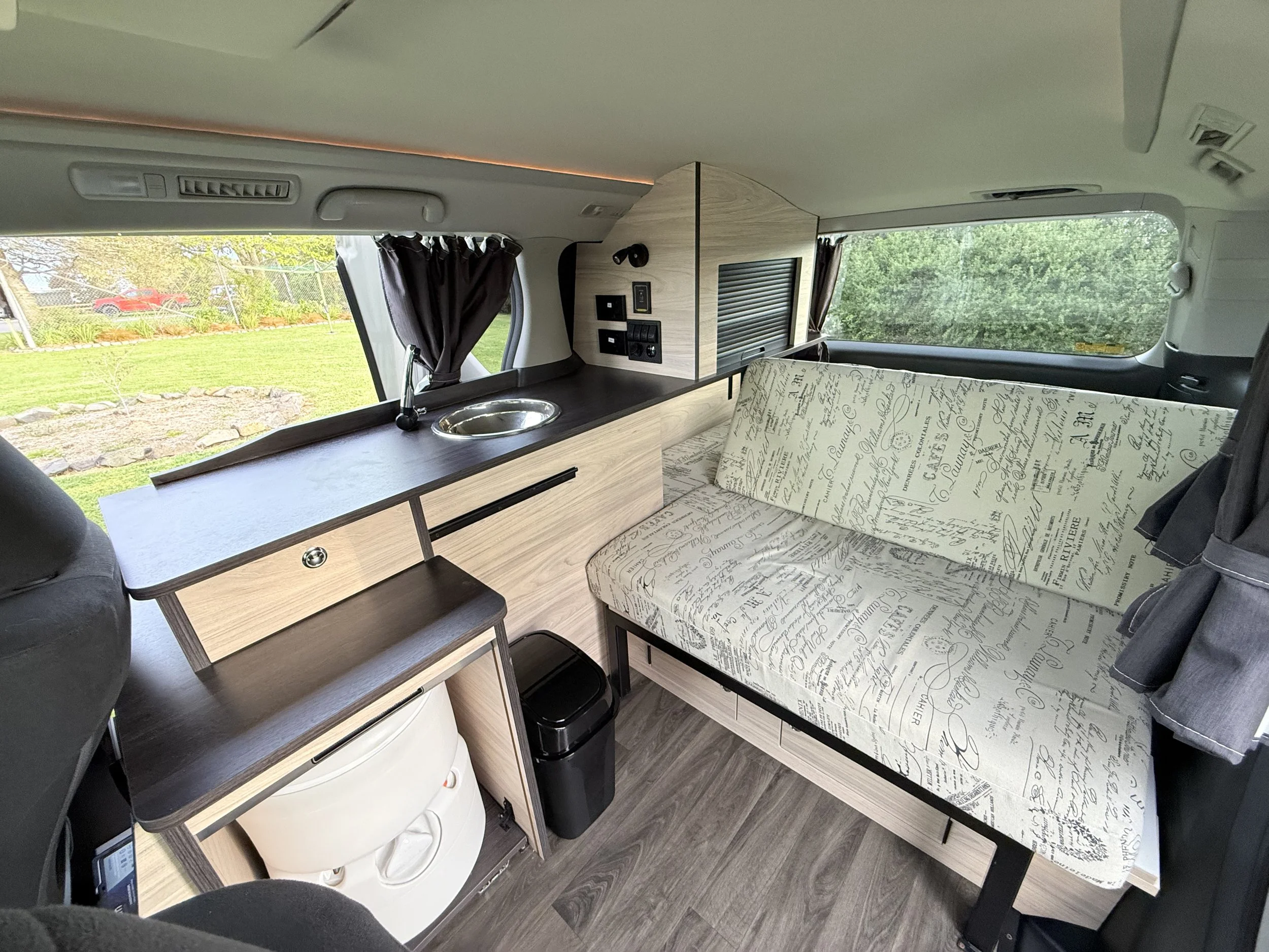 Interior of a camper van with a sofa, a small kitchen counter with a sink, a wooden partition, and windows with curtains, set in a grassy outdoor area.