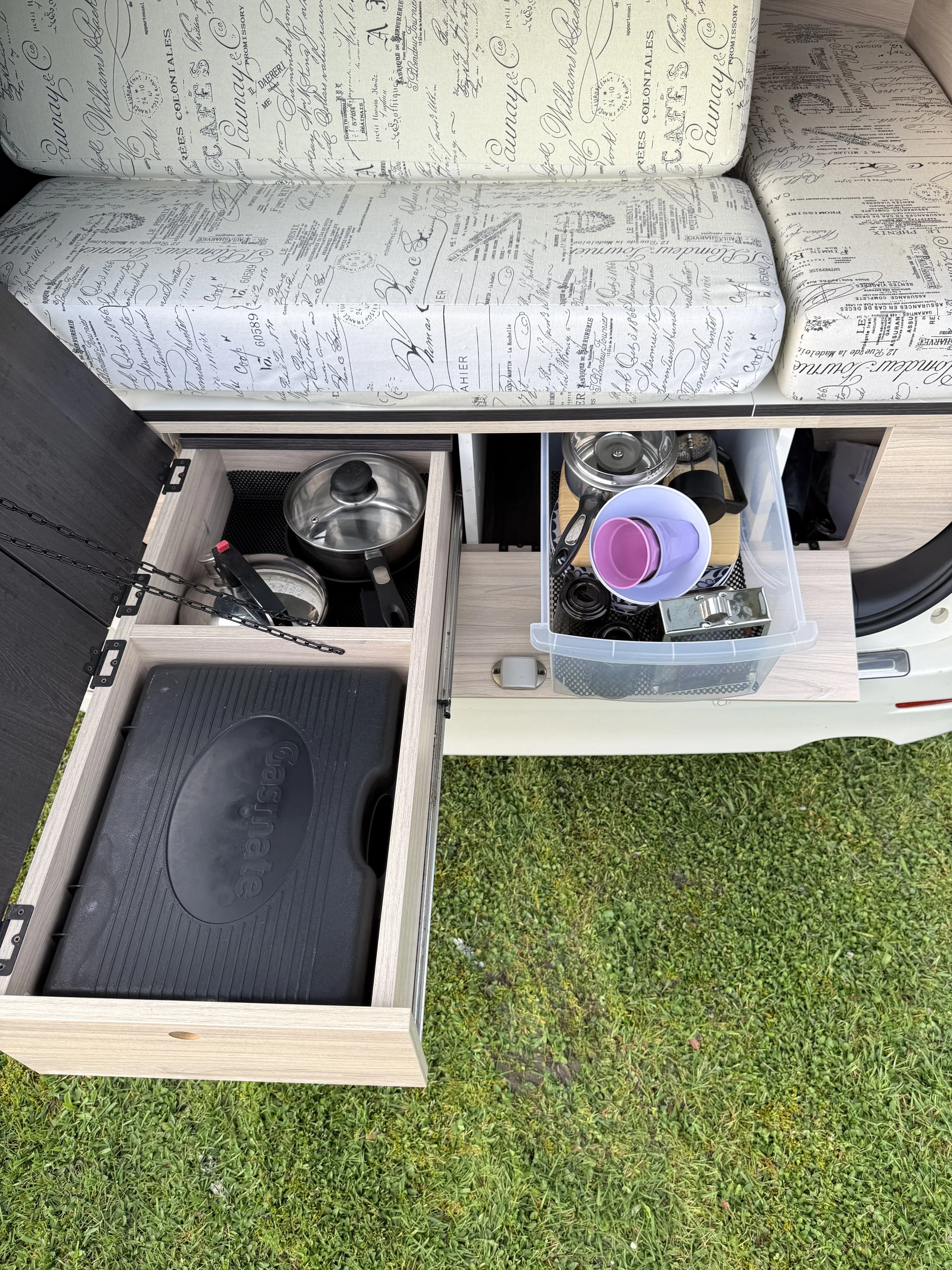 A small wooden outdoor kitchen area with a black grill, pots, and a plastic container with cups and kit. The area is on grass.