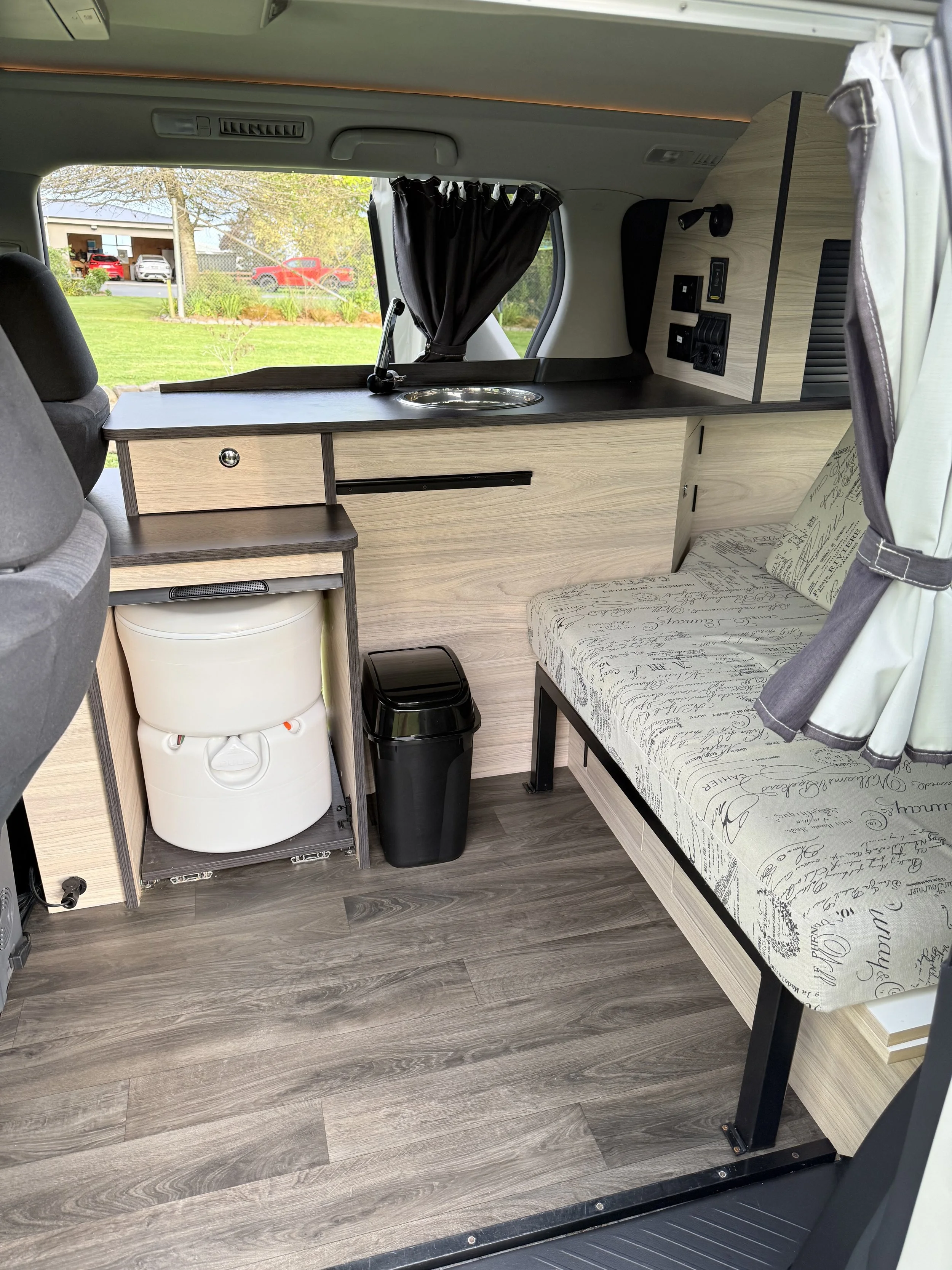 Interior of a camper van showing a sofa with printed fabric, a wooden kitchenette with a sink, electrical outlets, and a small refrigerator, set on a wood-like floor with a view of a garden outside.