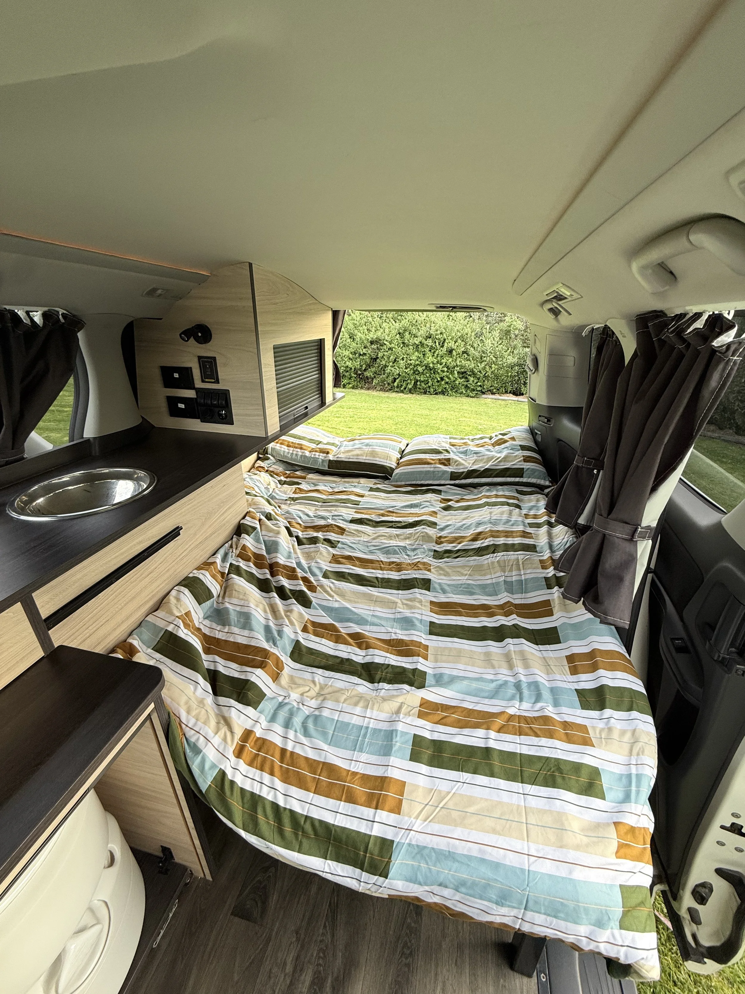 Inside a Go Beyond campervan, with a bed covered in a colorful striped blanket, small kitchenette with a sink, and a window with curtains showing green trees outside.