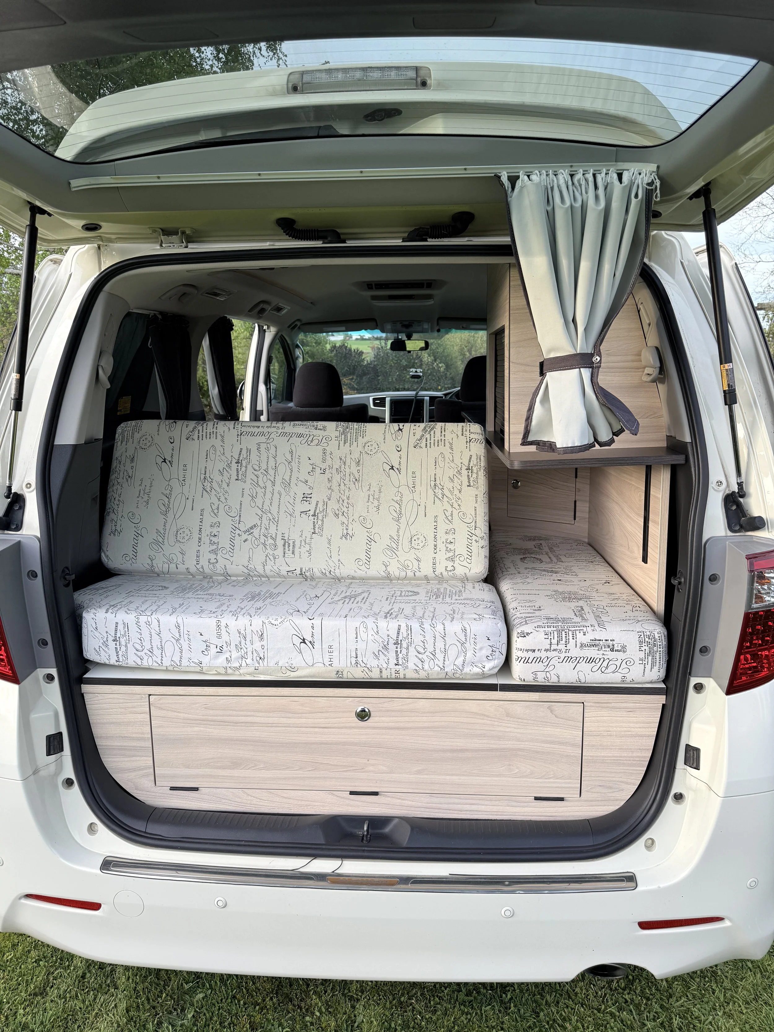 The rear of a camper van with the back door open, showing a convertible bench seat with upholstered fabric covered in script writing, a wooden storage cabinet, and curtains on the sides. The van is parked on grass with trees visible outside.