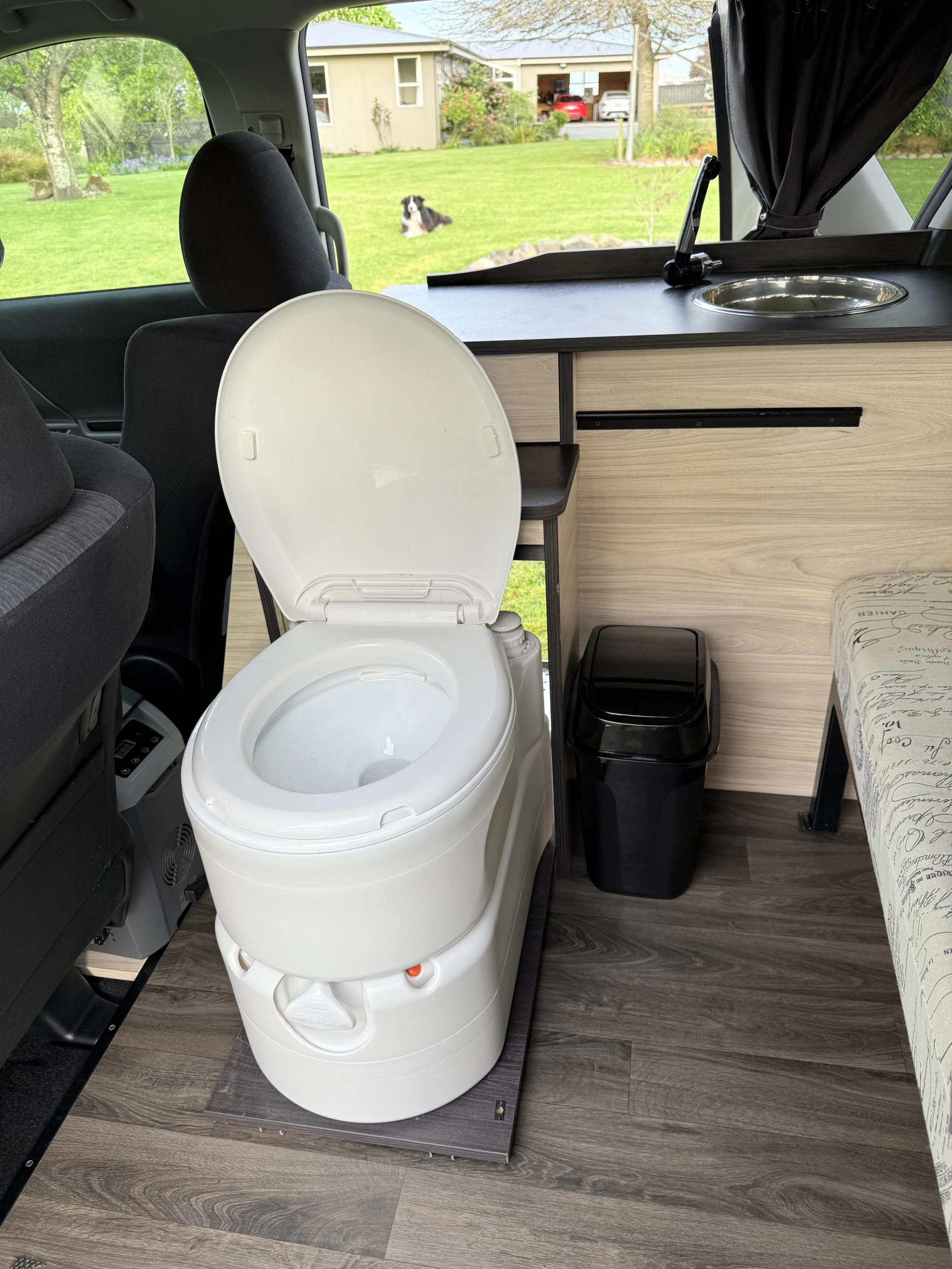 Inside a camper van, a white composting toilet is positioned on a dark wood platform near the front passenger seat, with a black trash bin beside it. A built-in wooden cabinet with a flat surface and a sink, covered with a black curtain on a rod, is 