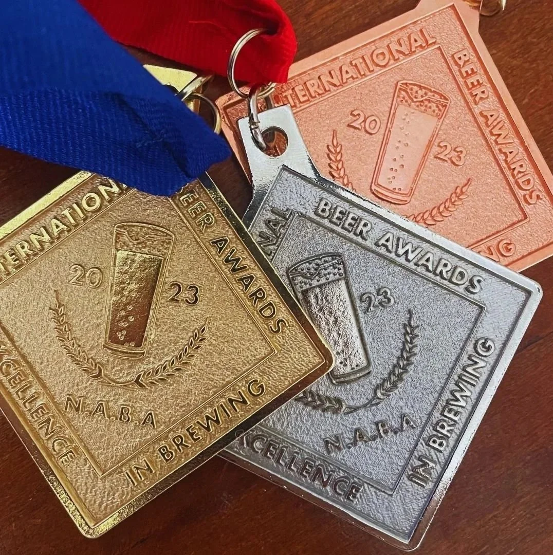 Three beer award medals in gold, silver, and bronze with ribbons, featuring a beer glass and laurel wreath, inscribed with 'International Beer Awards 2023 in Brewing' and 'NABA'.