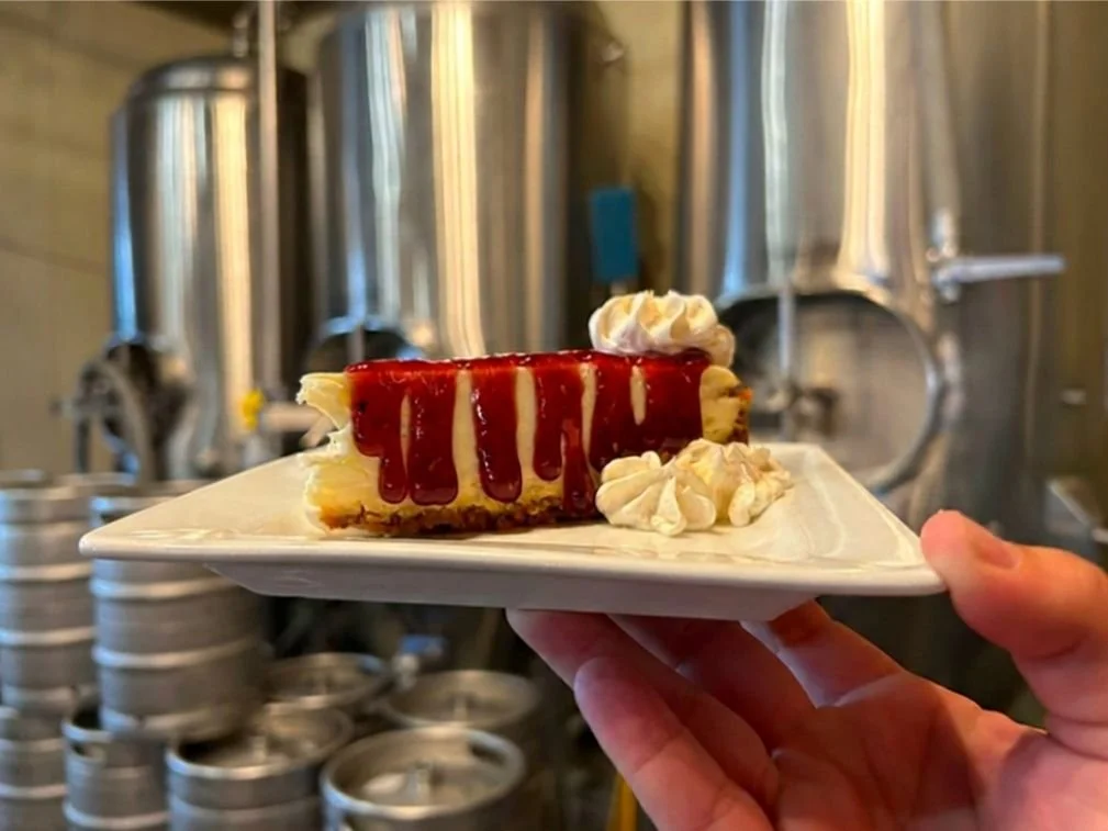 A slice of cherry cheesecake with whipped cream on top, served on a white plate, held by a person's hand with Bertram's stainless steel brewing equipment in the background.