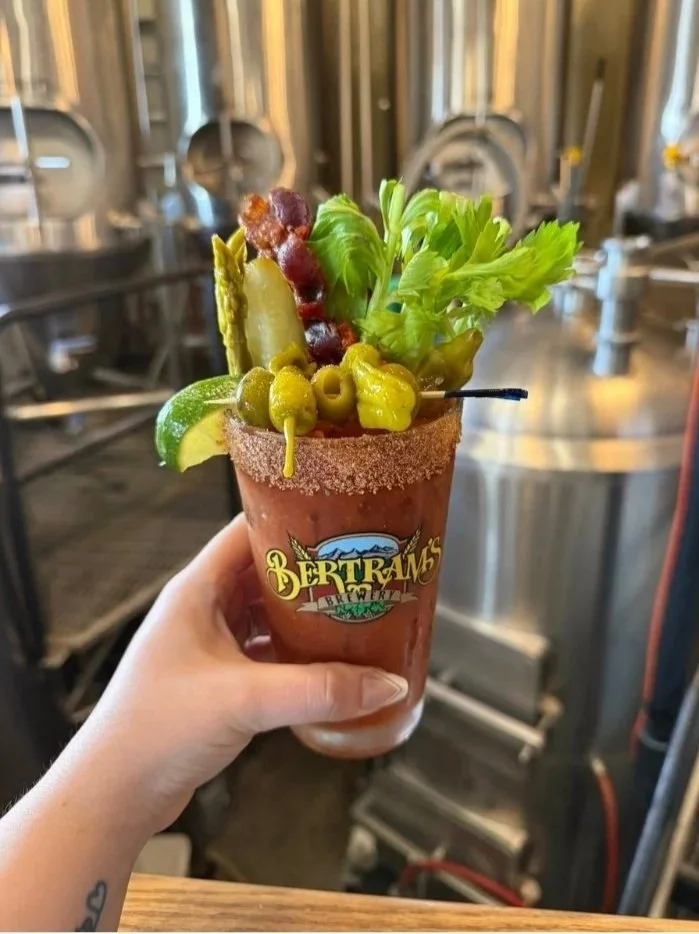 A Bloody Mary cocktail garnished with green olives, a lime wedge, celery, and pickles, served in a glass with a salted rim and Bertram's Brewery logo, in a brewery setting.