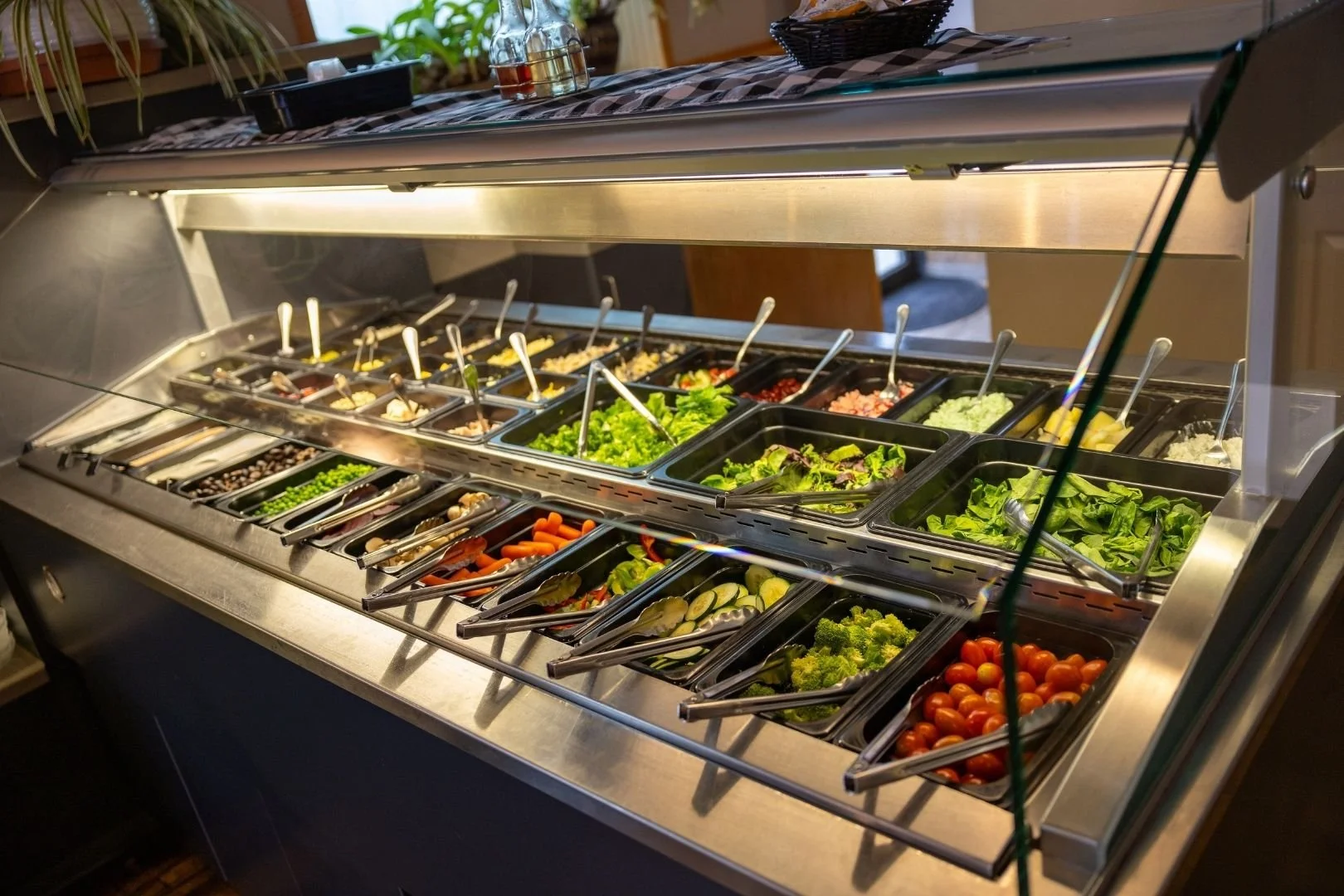 Salad bar with various fresh vegetables and toppings in black trays, covered by a glass guard in the Bertram's Brewery dining room.