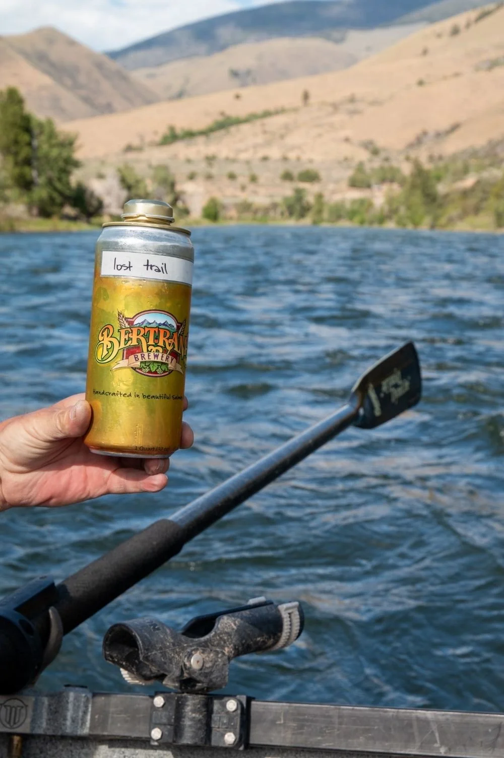 Fisherman holding a can of Bertram's 'Lost Trail IPA' in a driftboat on the Salmon River with mountains and trees in the background.