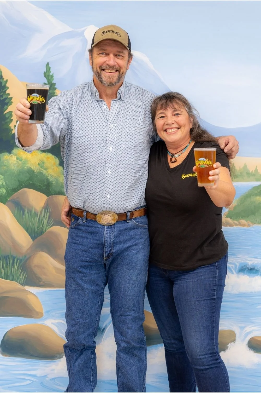 The husband and wife owners of Bertram's smiling and holding drinks in front of the painted mural in the Mountain Room event space.