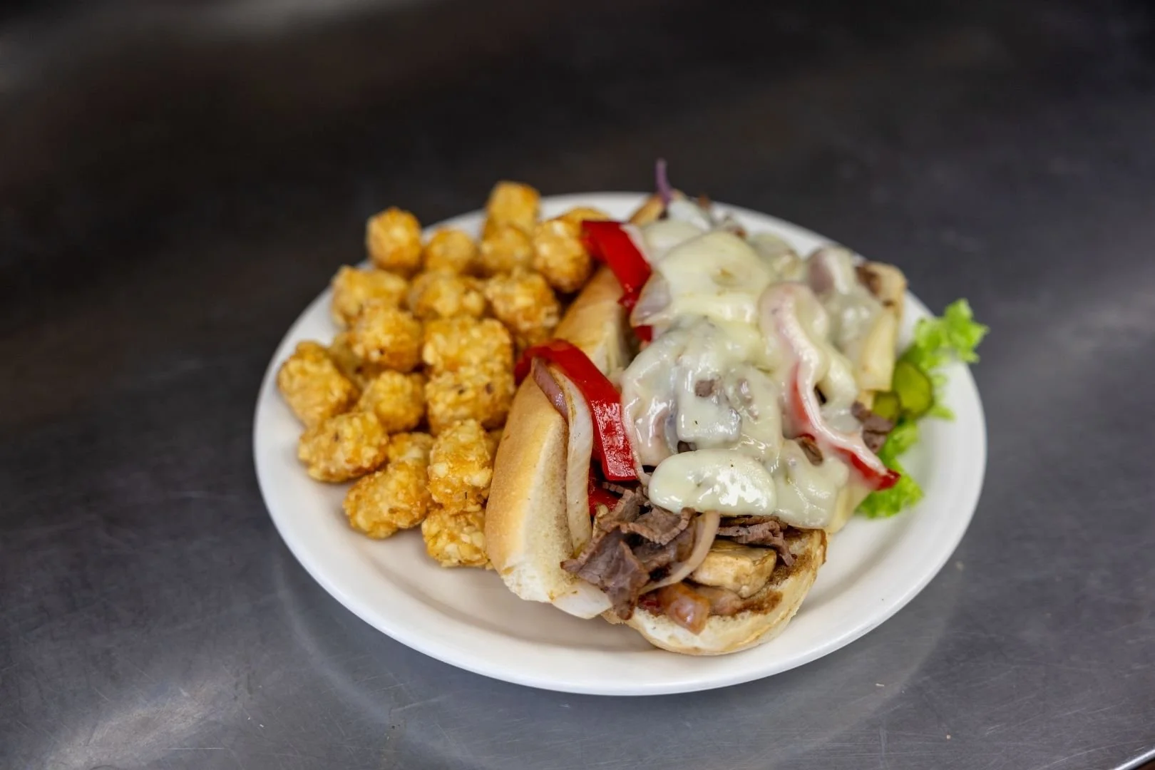 Plate with fresh tater tots and a hot sandwich with beef, red peppers, onions, lettuce, and melted cheese.