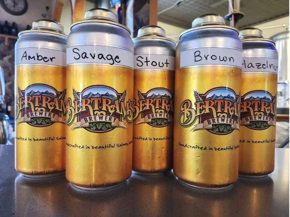 Five crowlers of beer with handwritten labels: Amber, Savage IPA, Sacagawea Stout, Brown Ale, Hazelnut, placed on the bar with brewery equipment in the background.