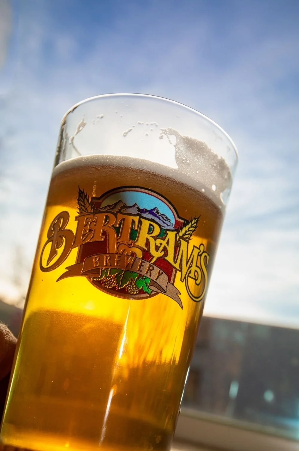 A glass of beer with a colorful Bertram's Salmon Valley Brewery logo, held up against a blue sky with light clouds.