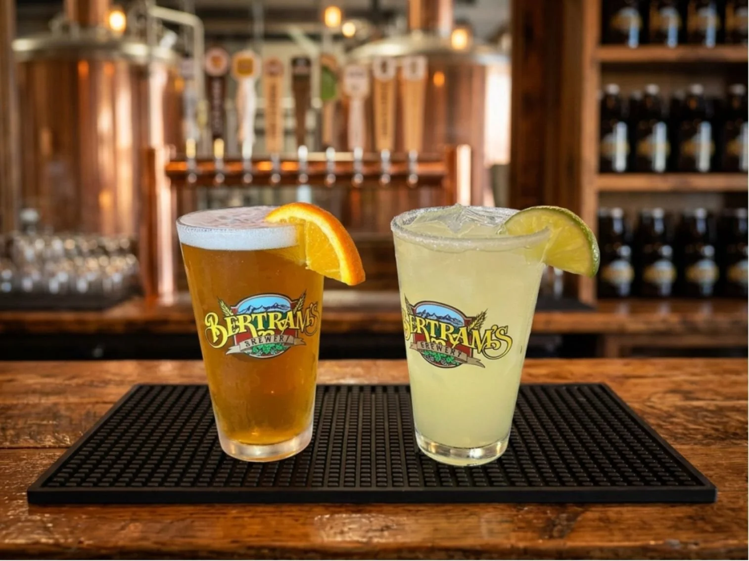 Beer and Margarita in Bertram's Brewery pint glasses, garnished with orange and lime slices, placed on a black mat on a wooden bar counter with a background of beer taps.