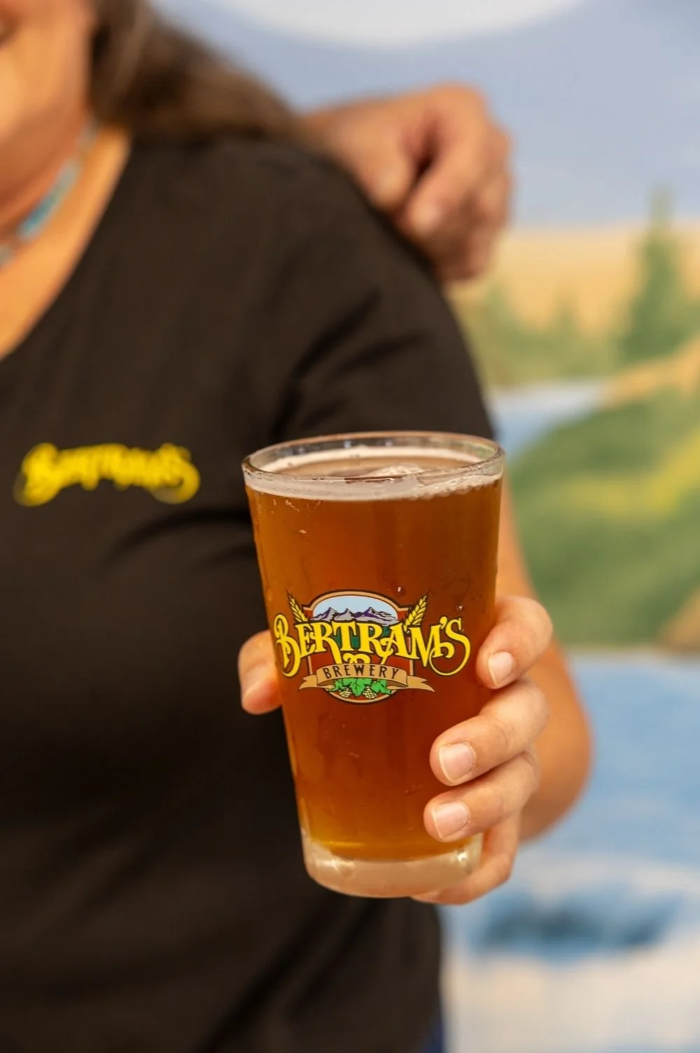 Melissa, the co-owner, holding a pint glass of amber beer with the Bertram's Brewery logo, background of a scenic mural of the Rocky Mountains.