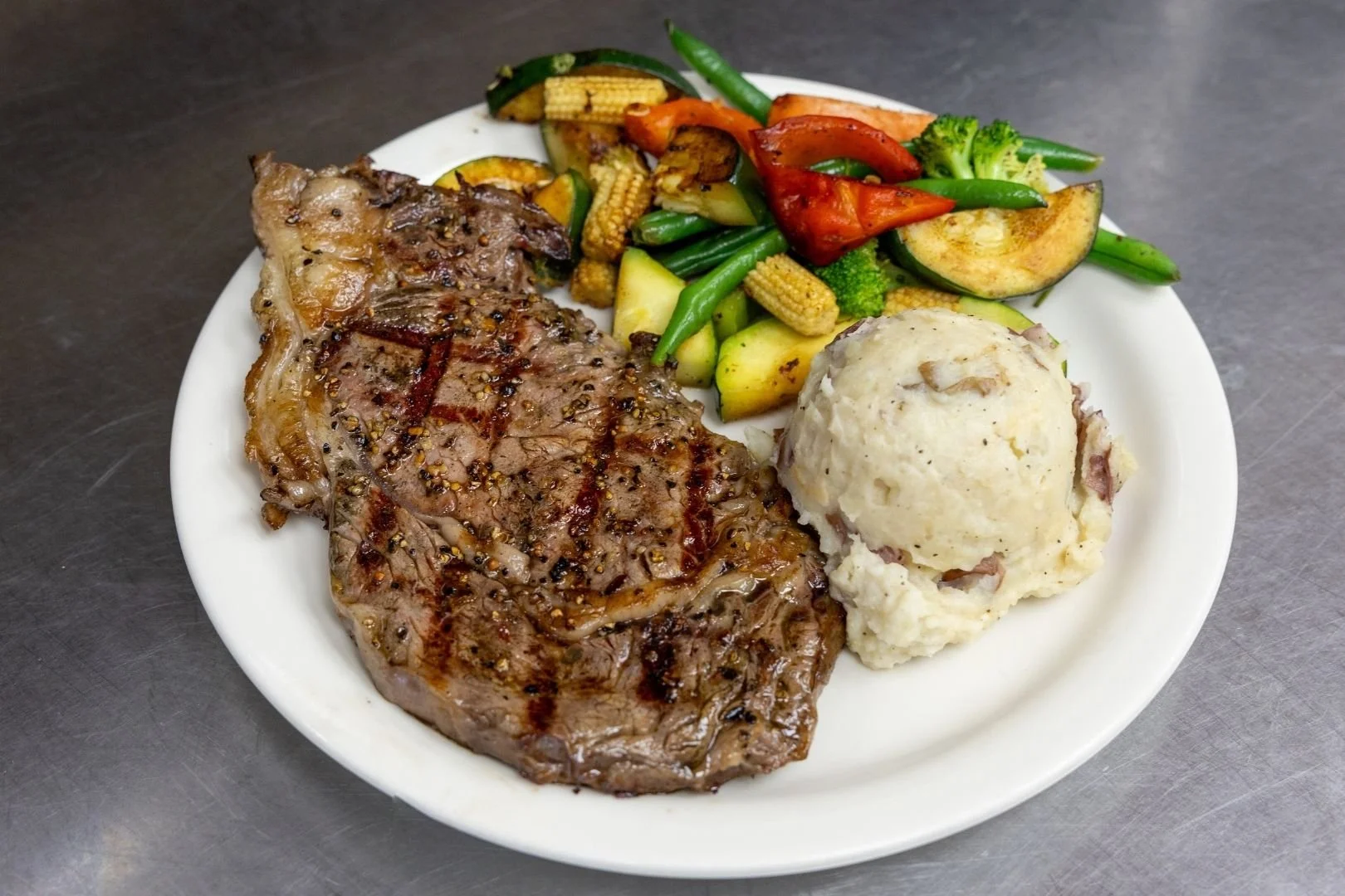 Plate with grilled steak, mashed potatoes, and mixed grilled vegetables including zucchini, carrots, broccoli, green beans, and baby corn.
