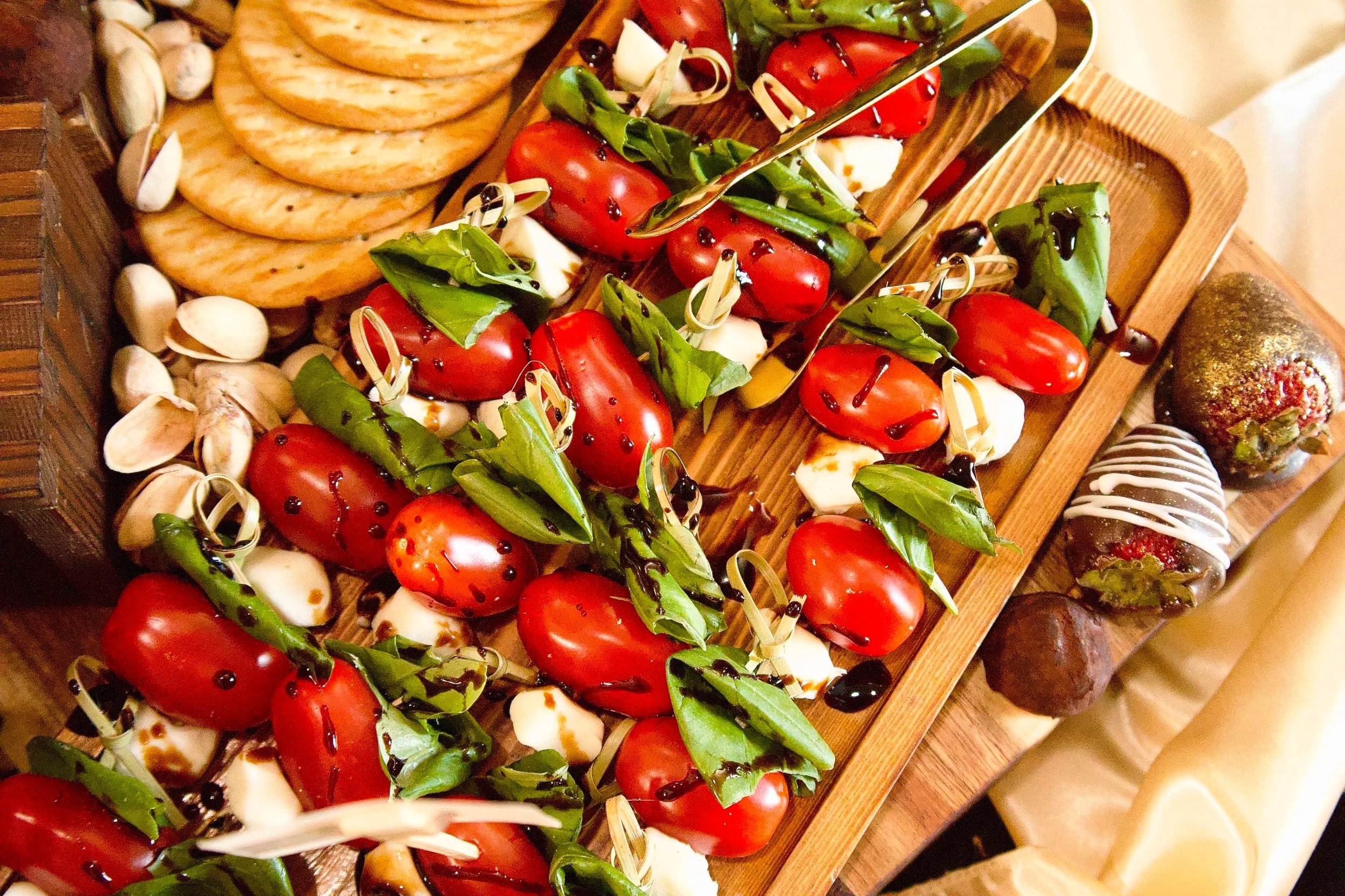 A wooden tray with caprese skewers containing cherry tomatoes, fresh basil, mozzarella, and balsamic glaze, arranged with some crackers and nuts nearby.