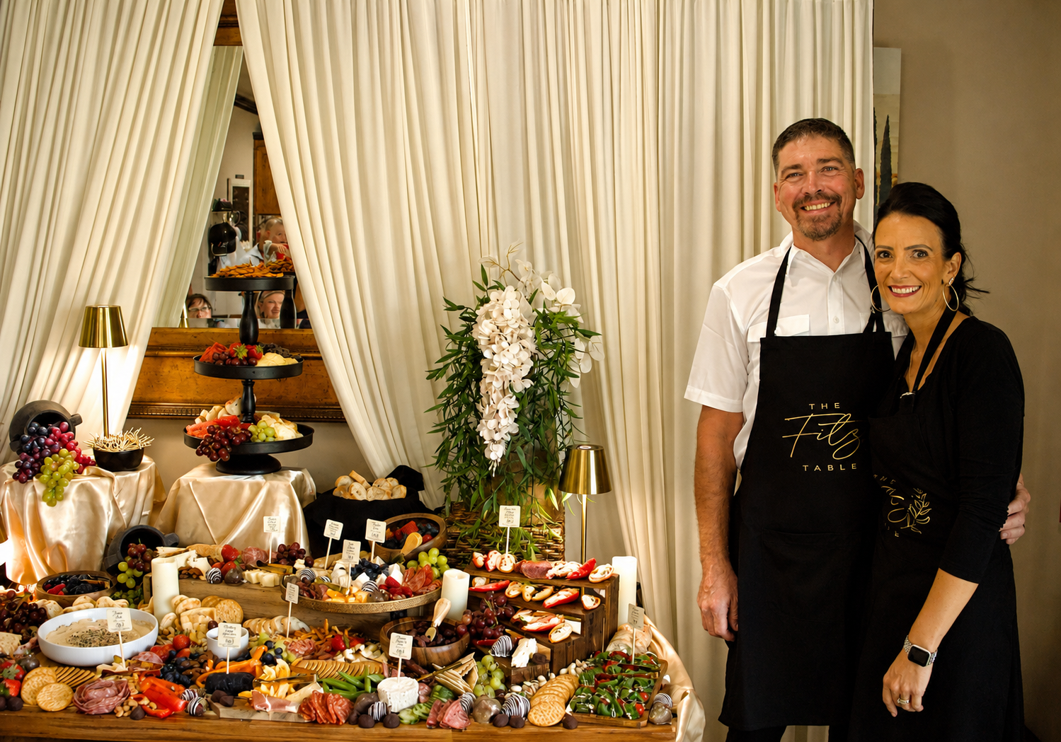 Two people standing in front of cheese and charcuterie platters with fruits, crackers, and dips, at a social event with cream curtains and a mirror in the background.