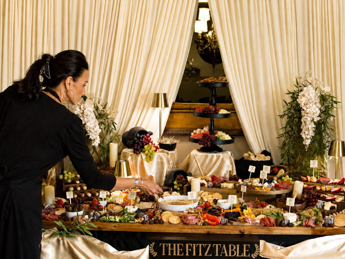 A woman arranging a cheese and charcuterie platter at a buffet table decorated with candles and floral arrangements, with a sign reading 'The Fitz Table'.