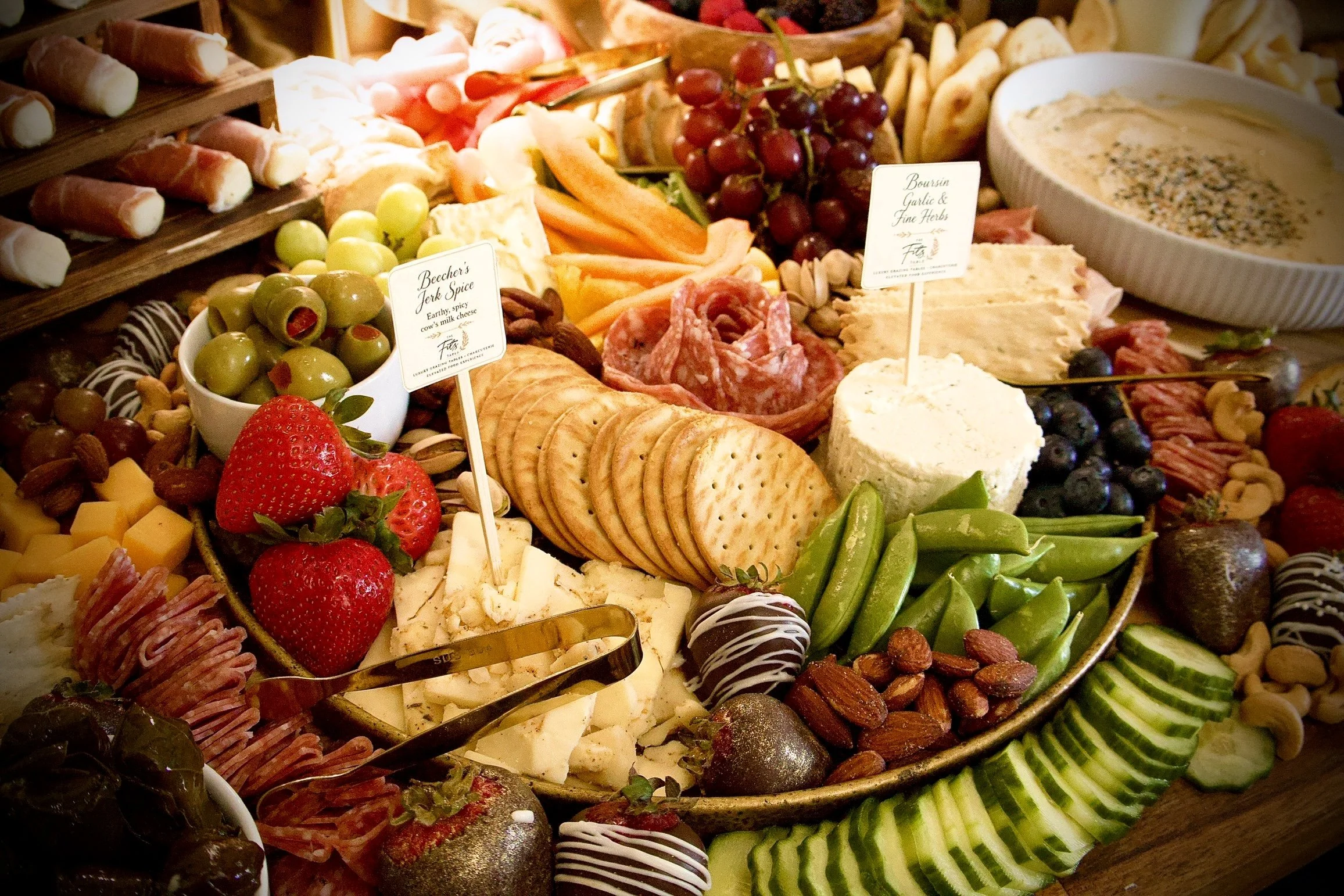 A large platter of assorted cheeses, fruits, nuts, crackers, meats, and chocolate truffles, with small labels indicating different items.