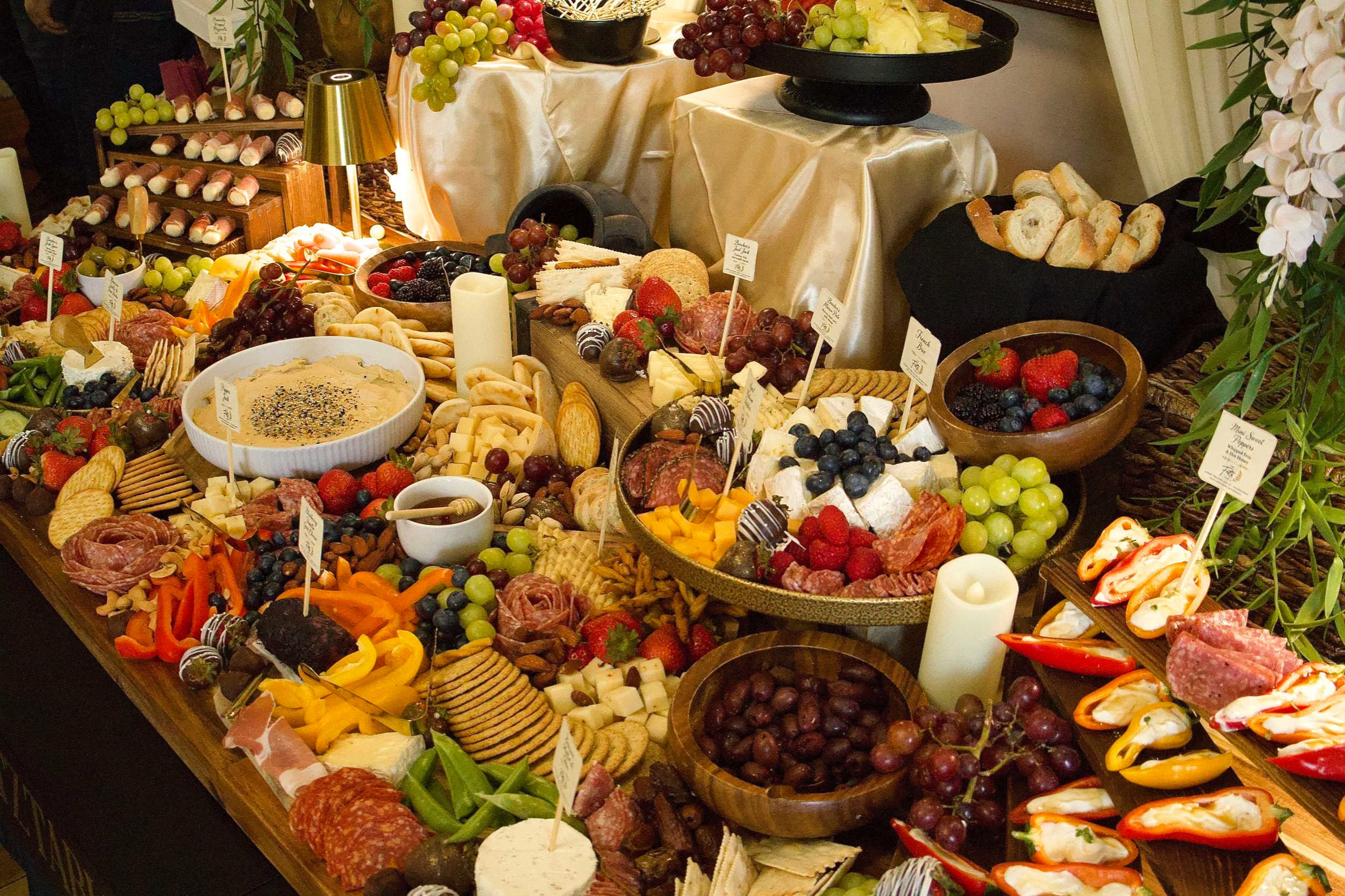A large spread of assorted cheeses, fresh fruits, meats, crackers, and dips on a buffet table at an event, decorated with greenery and candles.