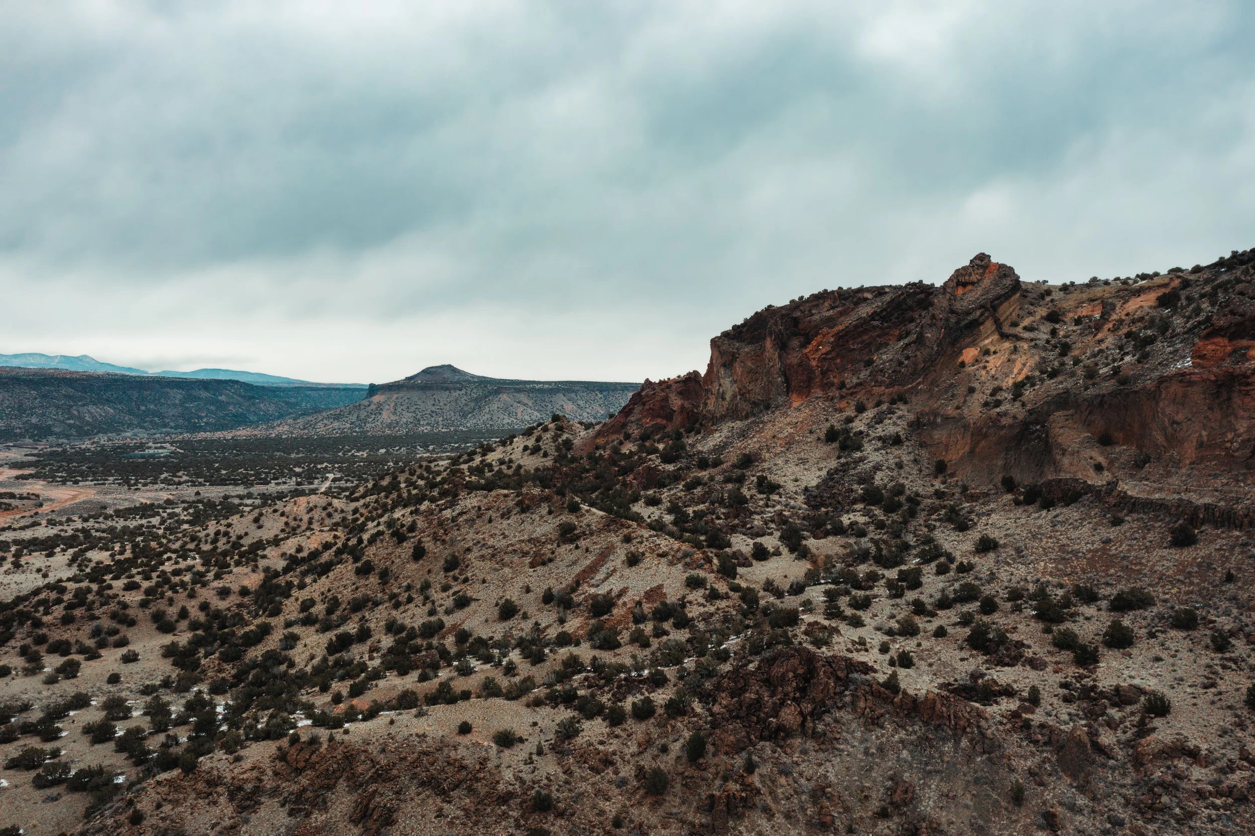 A wide desert landscape with rocky formations, sparse vegetation, and an overcast sky.