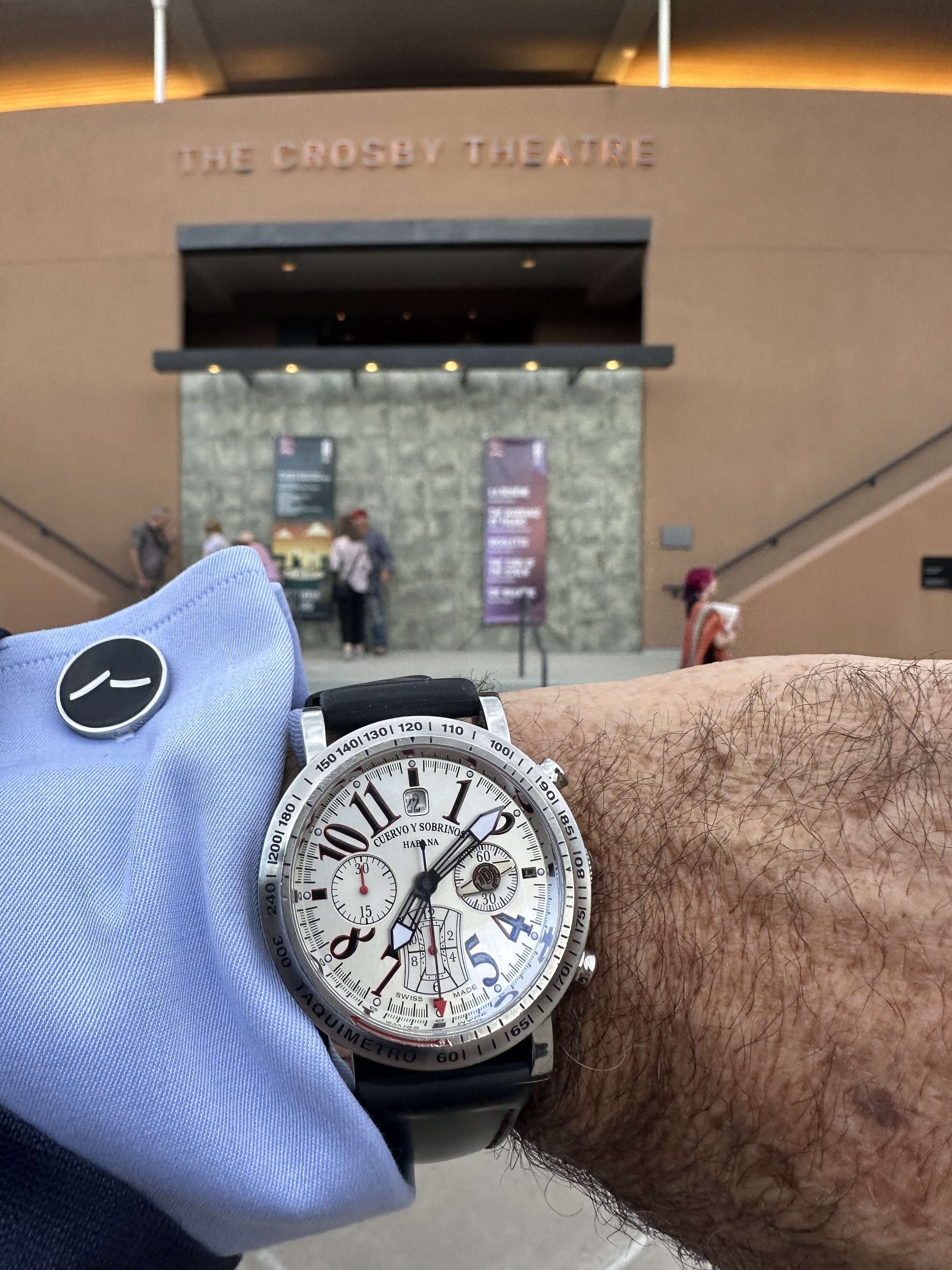 A person's wrist wearing a large, white chronograph watch with black strap and red accents, on a hairy arm, resting on a surface in front of a theater entrance. The background shows a sign for The Crossey Theatre with a stone wall and a few people standing underneath.