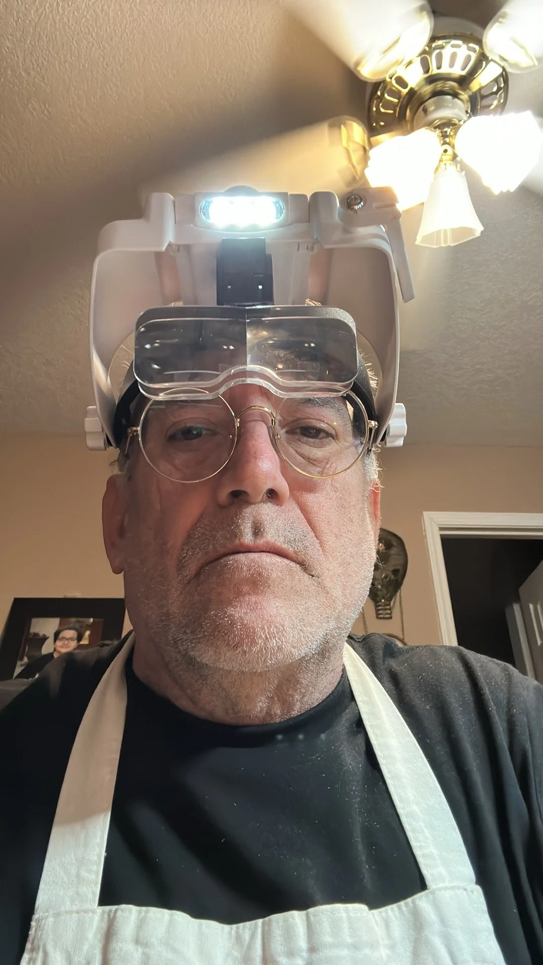 An older man with glasses wearing a dental surgeon's headlamp and a white apron, standing in a room with a ceiling fan and a decorated wall in the background.