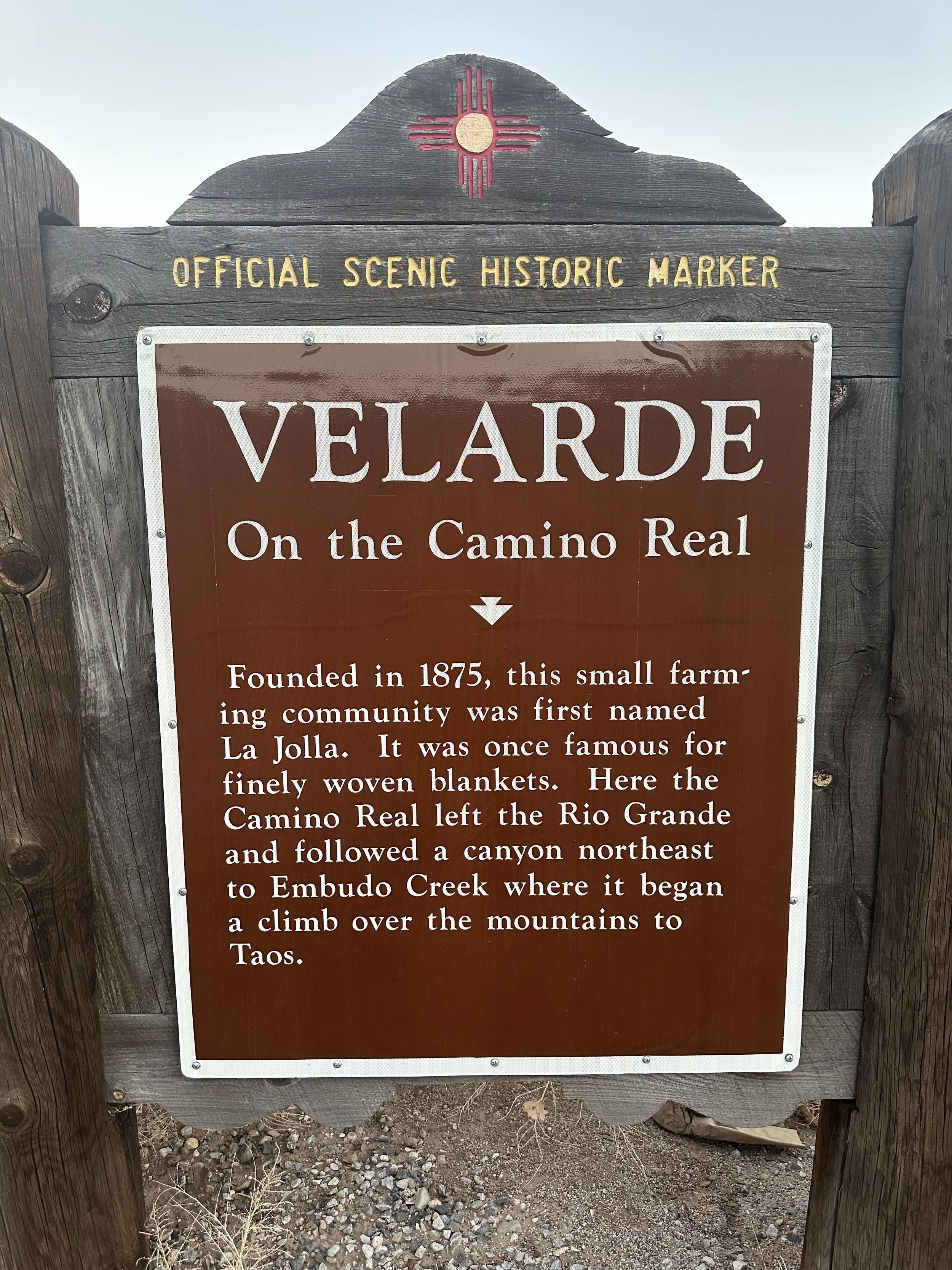 A wooden sign with a historical marker about Velarde, located on the Camino Real. The sign mentions Velarde was founded in 1875, initially called La Jolla, and describes its location and significance in connecting to Embudo Creek.