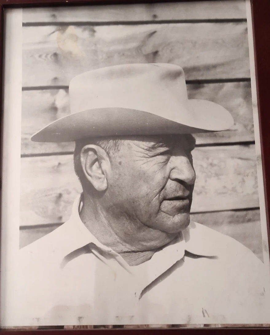 A black-and-white photograph of an older man wearing a wide-brimmed hat, with a background of wooden planks.