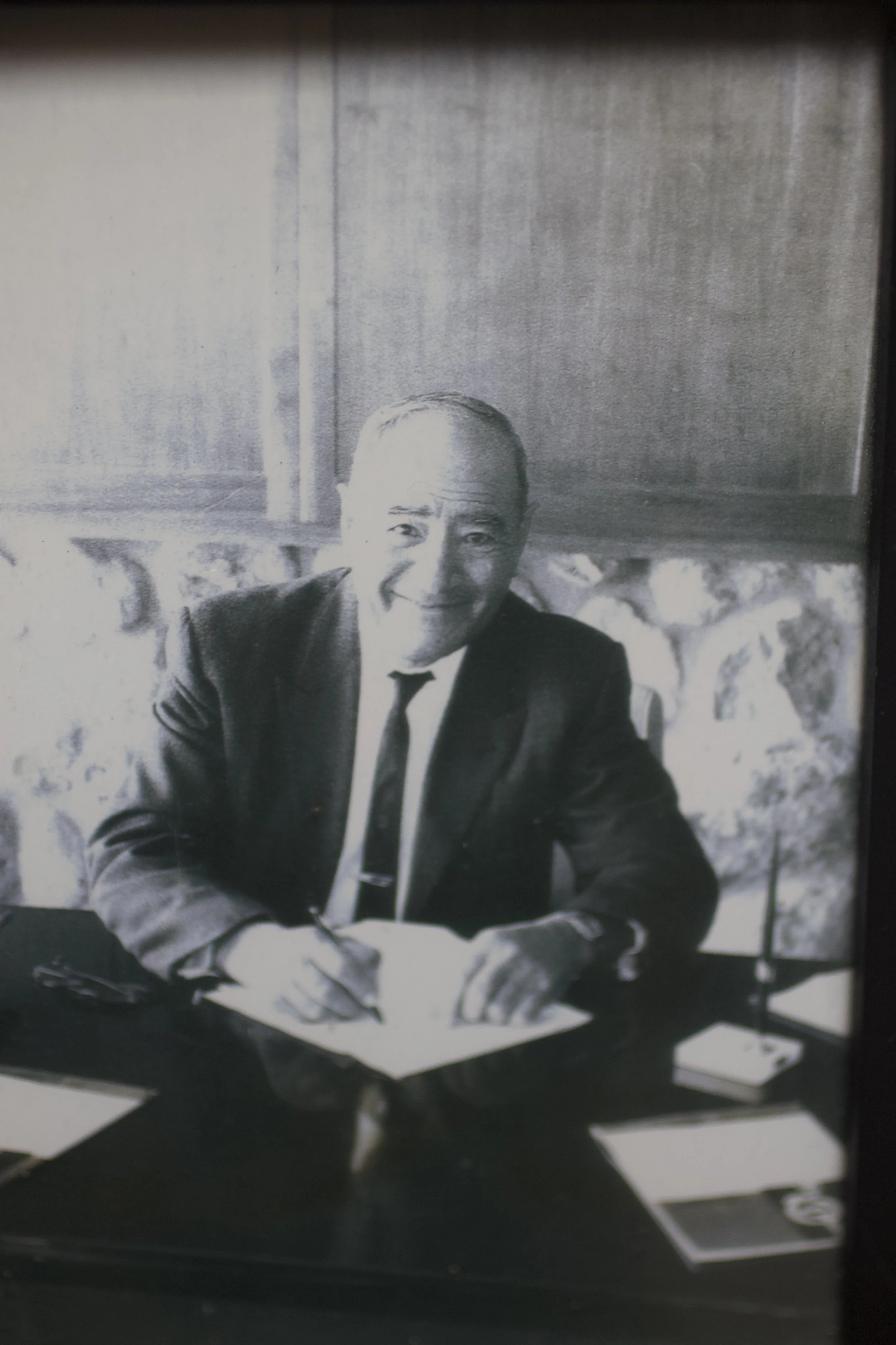 Black and white photograph of a smiling man in a suit sitting at a table with papers in front of him.