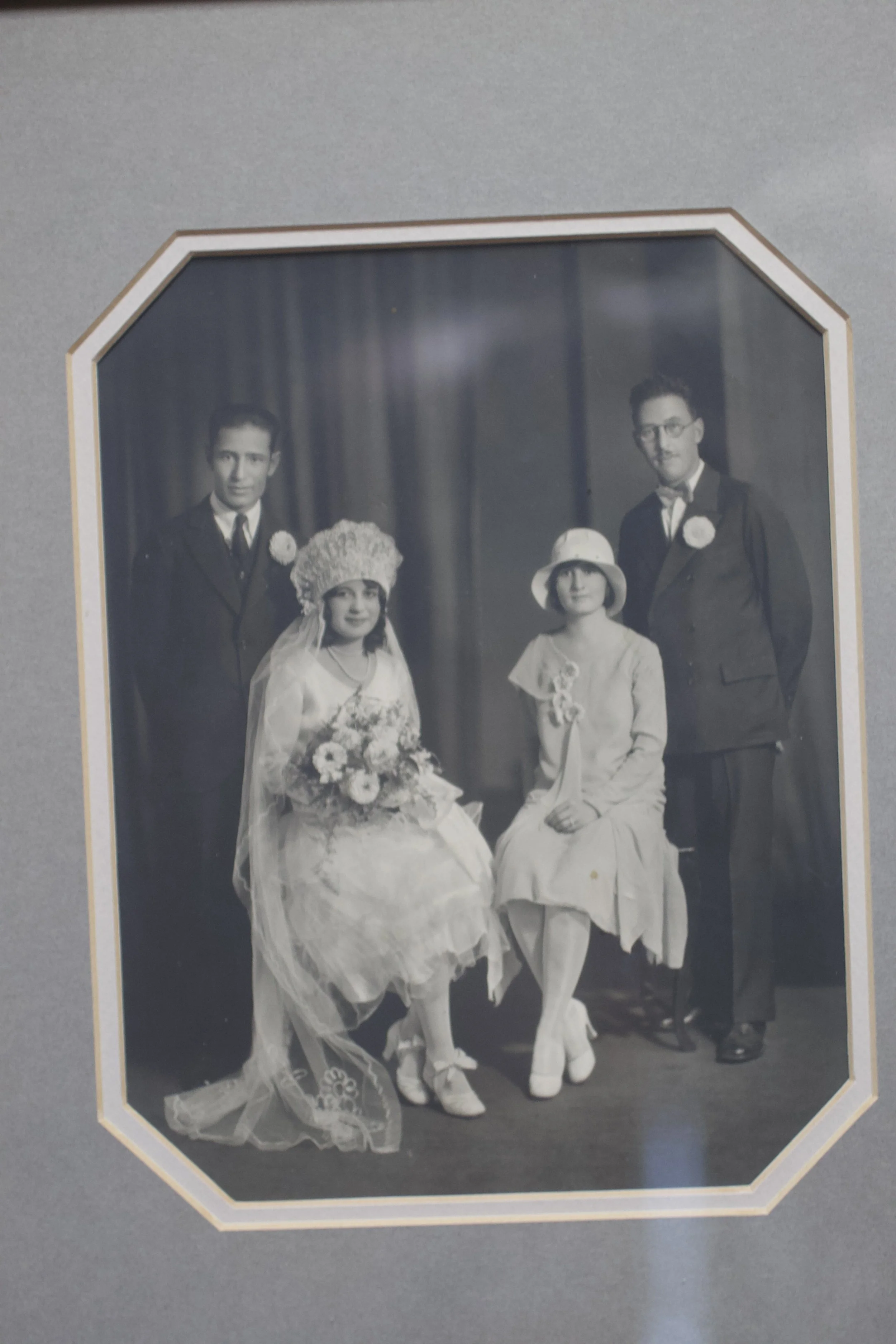 A vintage black and white wedding photograph showing four people: a seated bride in a lace wedding dress with bouquet, a standing bride in a light-colored dress and hat, and two men in suits with boutonnieres. The photo has an octagonal border.