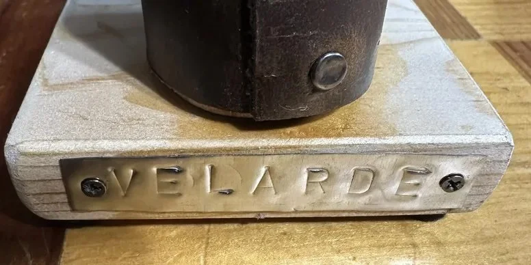 Close-up of a wooden base with a metal nameplate that reads 'VELARDE' attached by two screws, and a black object on top of the base.