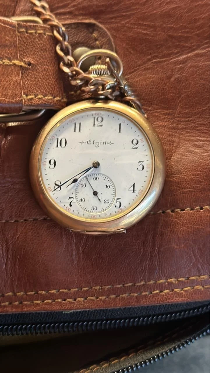A vintage Elgin pocket watch with a white dial, black Arabic numerals, and a smaller seconds subdial at the 6 o'clock position. The watch has a gold case and is attached to a metal chain, resting on a brown leather surface.