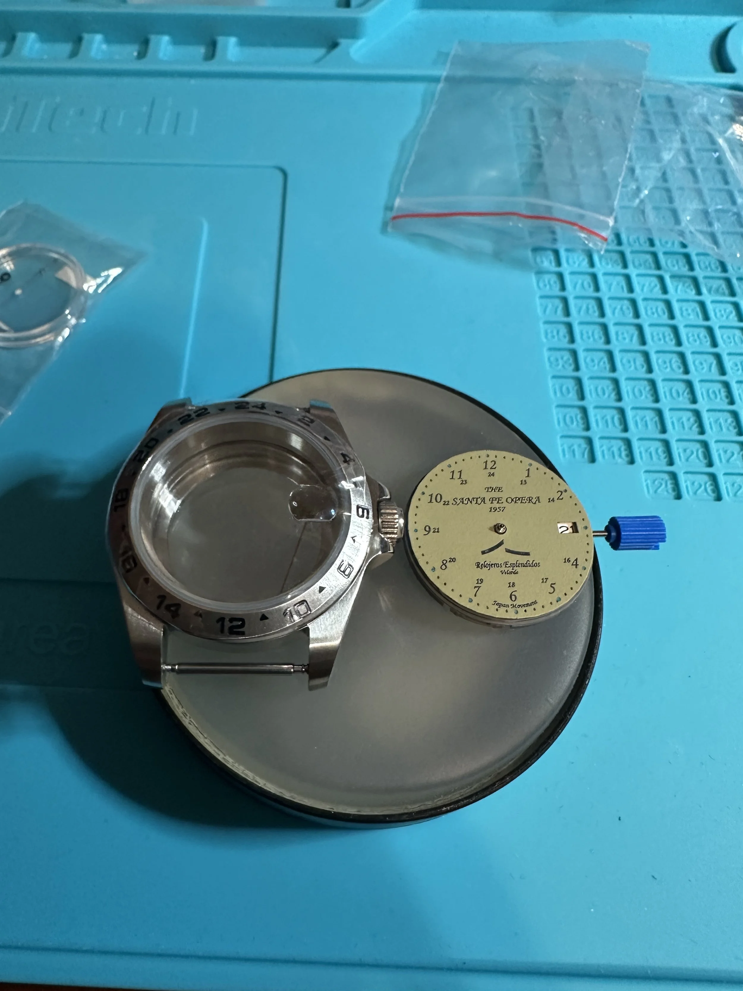 A wristwatch with a metal band and a black bezel, placed on a blue surface with a clock face inscribed with "SANTA FE OPERA 1957" and other markings. There is a small clear plastic container and a piece of clear tape nearby.