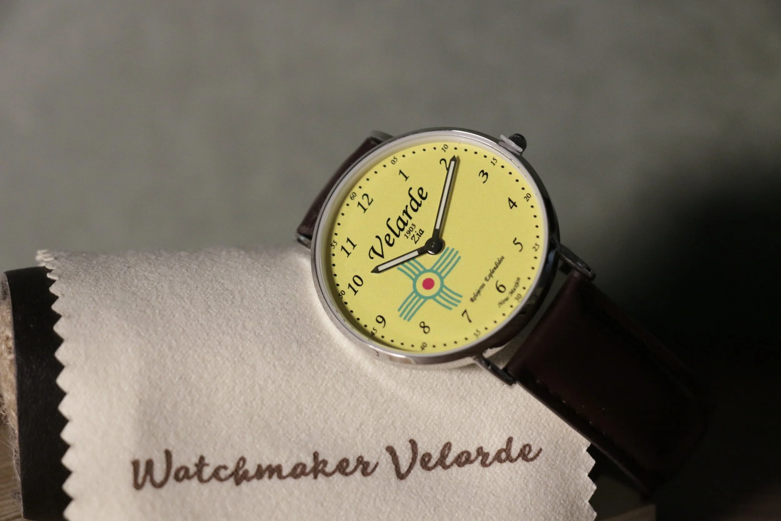 A yellow watch with black numbers and a brown leather strap, displaying the time around 10:09, placed on a white textured surface.