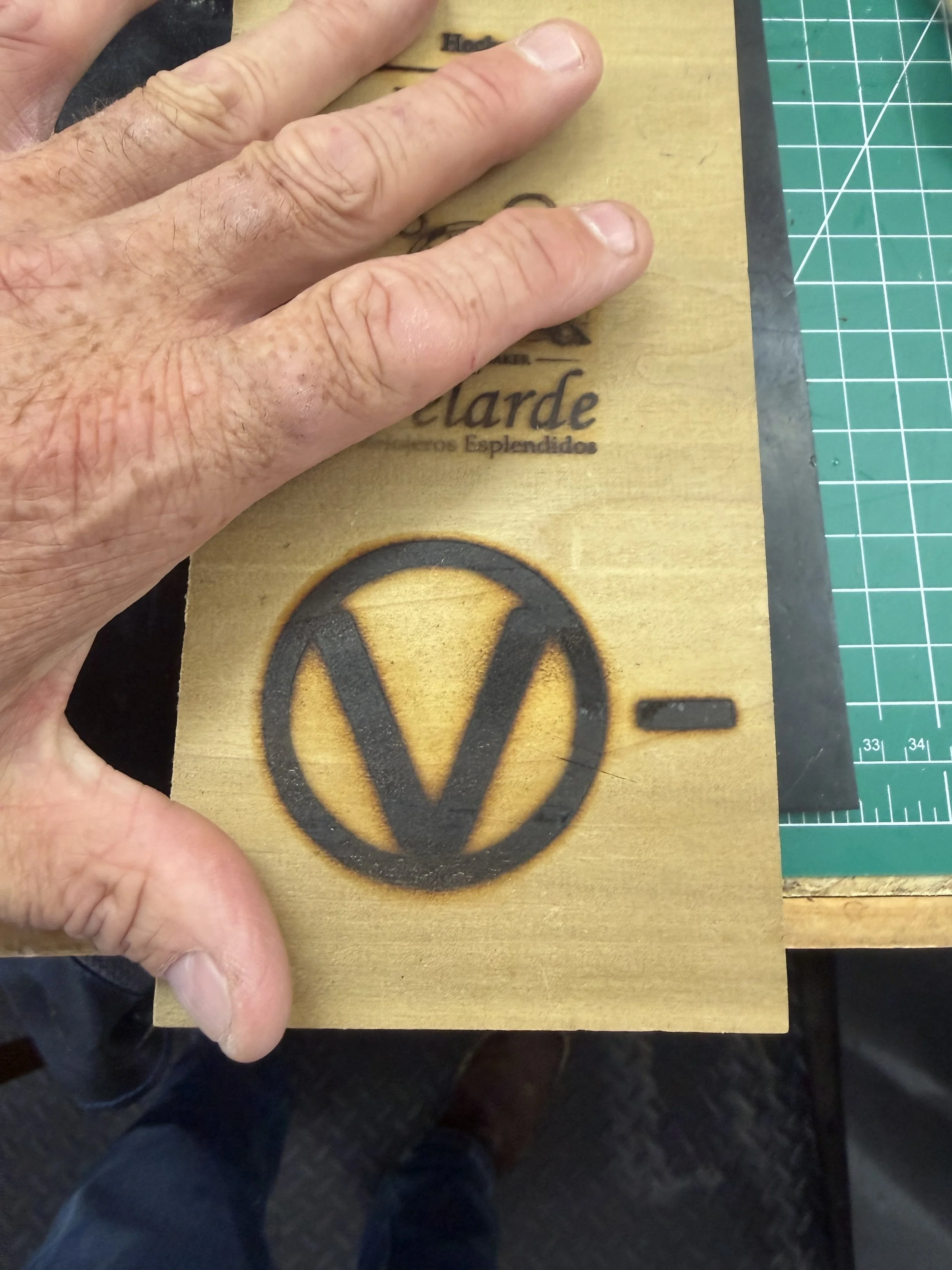 A hand with hairy skin and short nails holding a wooden plank with a black Volkswagen logo and partially visible text on it, on a green cutting mat.