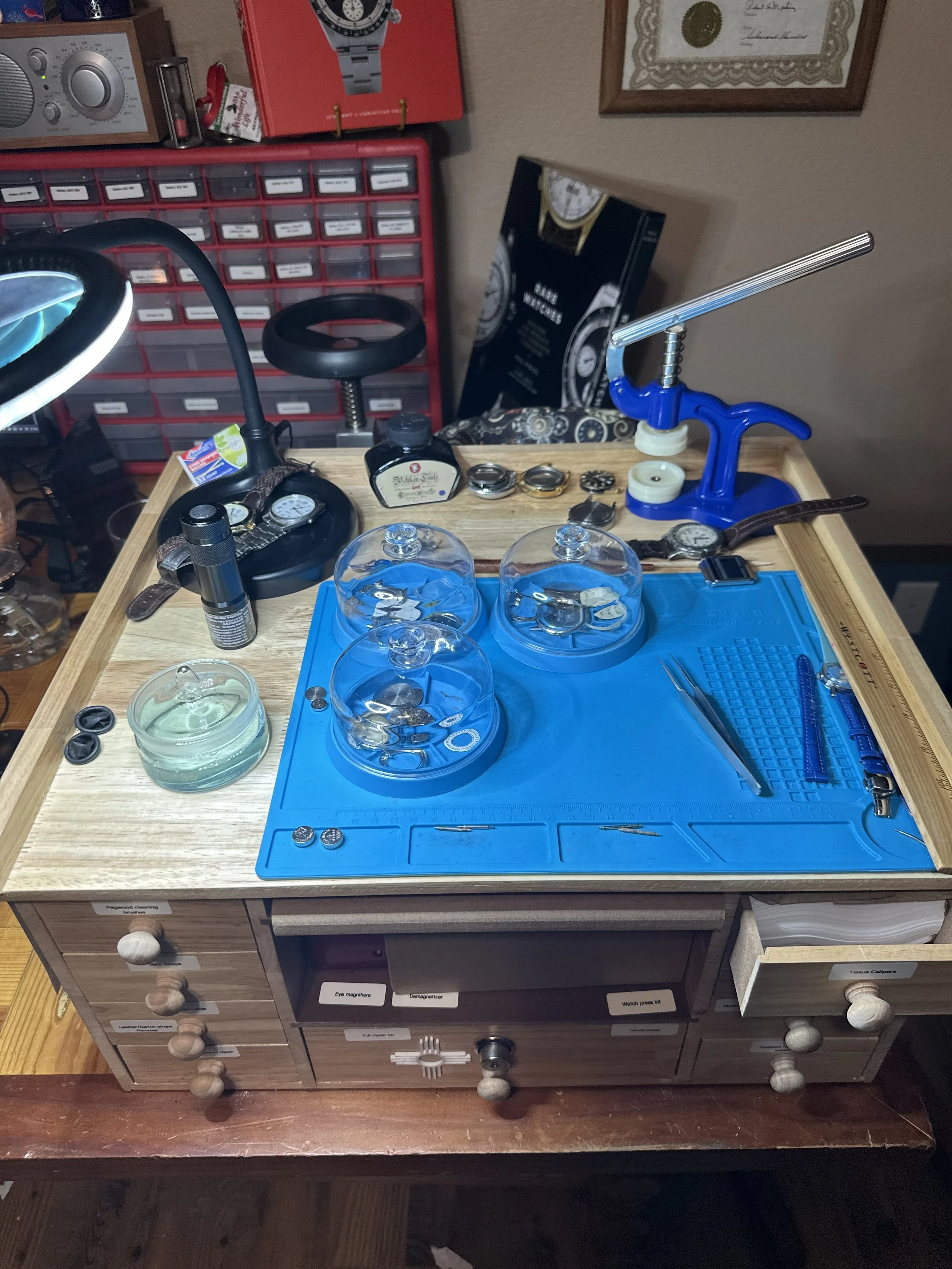 A wooden desk with a light blue tool mat, displaying various watch parts including watch faces, bands, and glasses, along with tools and bottles of oils. Behind the desk, a red storage cabinet filled with small labeled compartments and framed certificates or awards are visible.