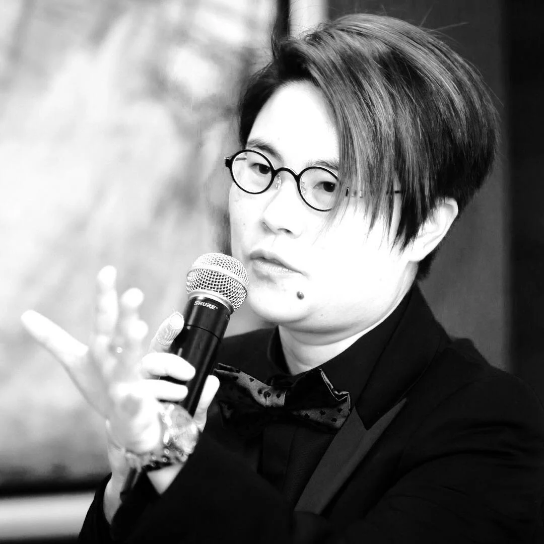 Vince Chan speaks into a microphone in a black-and-white event photo, dressed in a suit and bow tie with a focused expression.