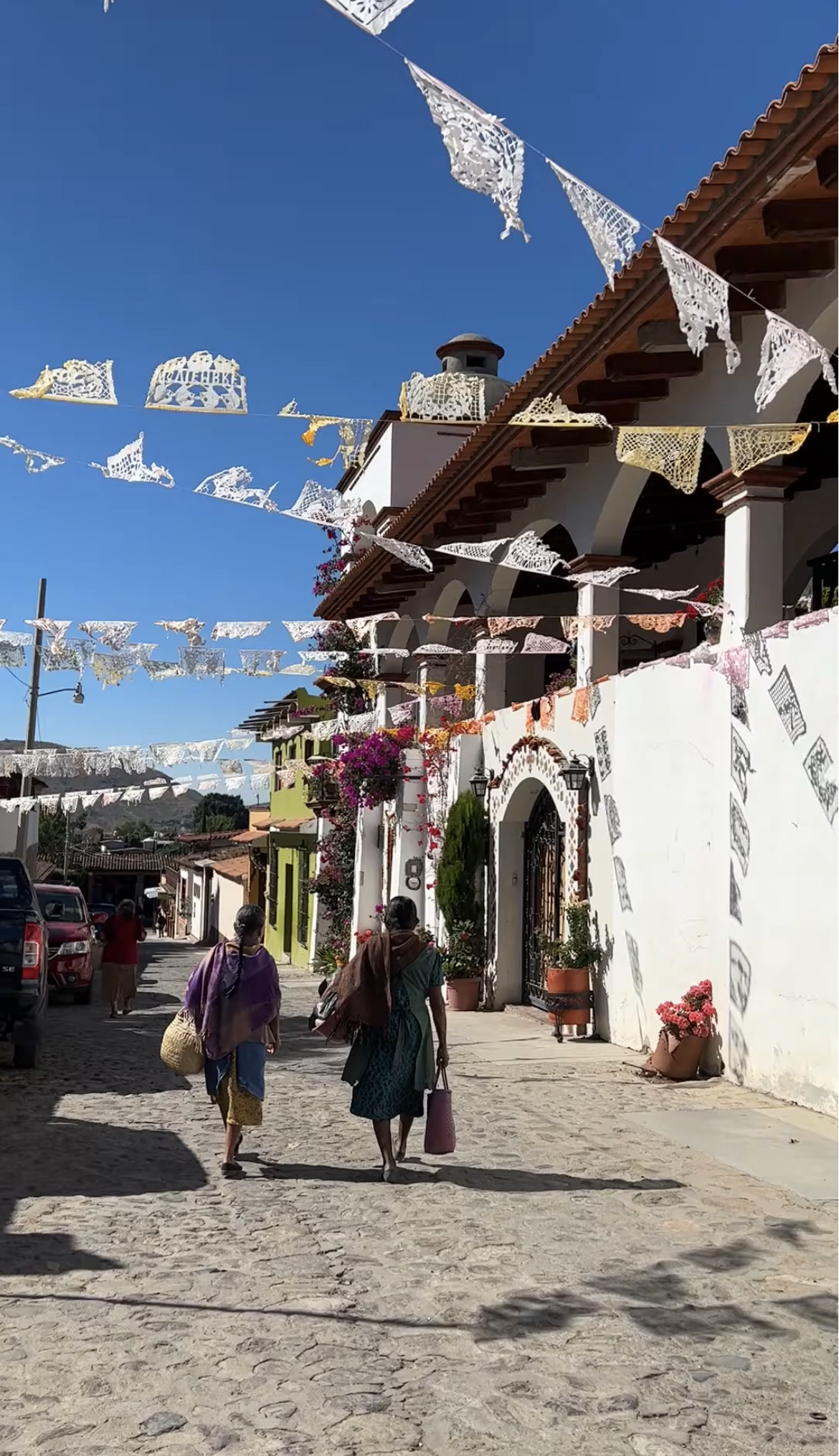 A street scene in a Mexican town with white buildings, cobblestone pavement, and colorful flowers. People are walking, and decorative paper flags are hanging across the street under a clear blue sky.