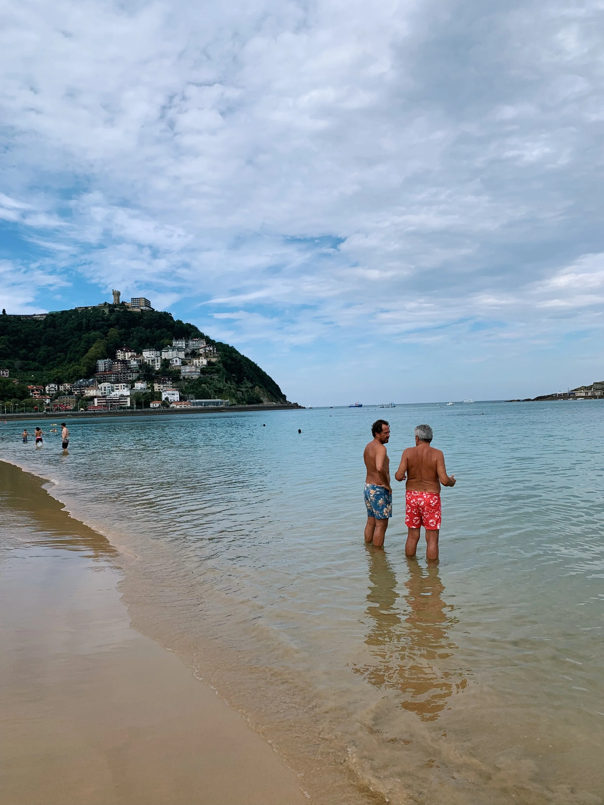 Two men standing in shallow water at the beach having a conversation, with other people swimming and playing in the water nearby. A hillside with houses and a tower is visible in the background under a partly cloudy sky.